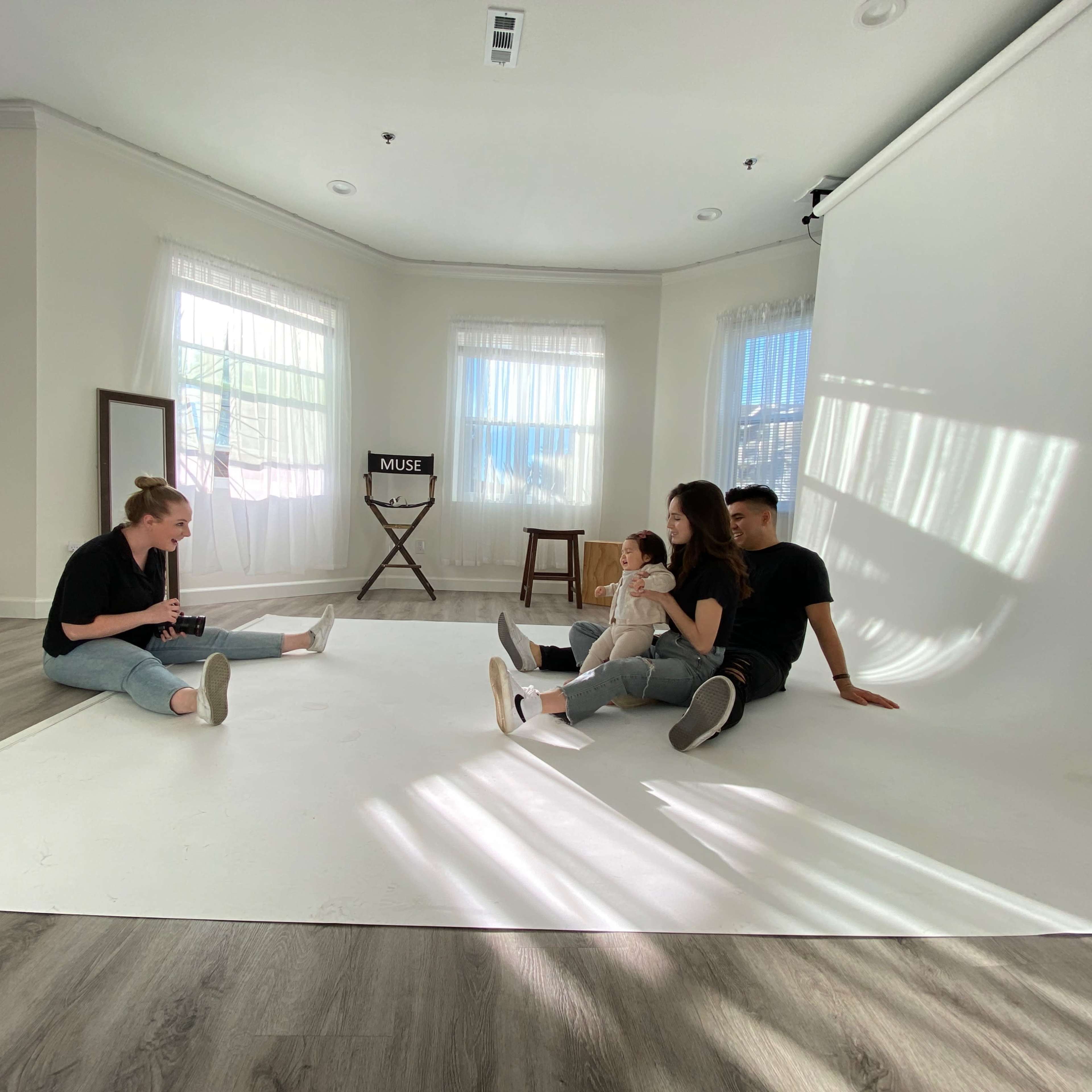 A group of four people sits on a white floor in a bright studio with large windows and a backdrop.