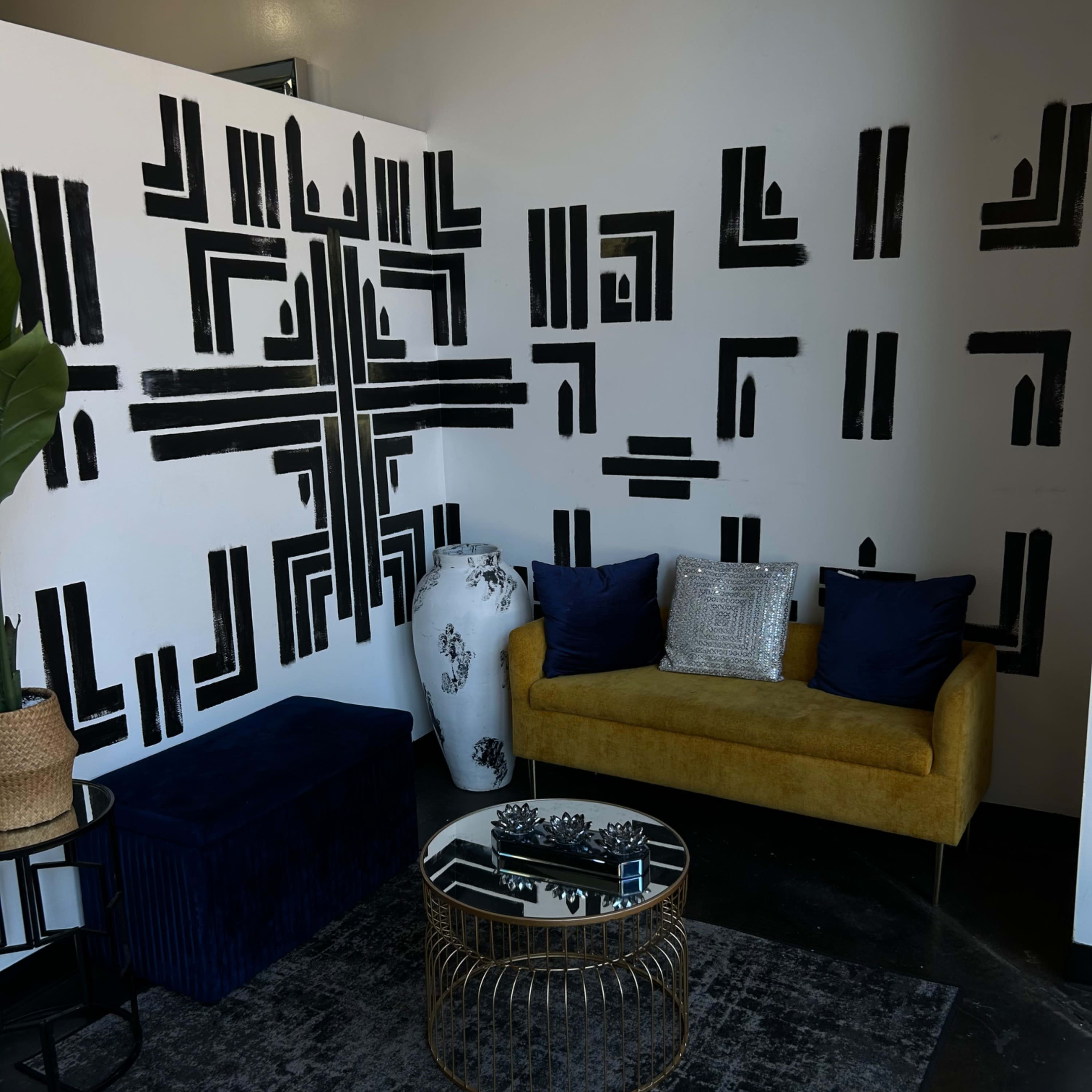 A modern seating area with a yellow sofa, a black patterned wall, and decorative elements including a glass coffee table and a large plant.