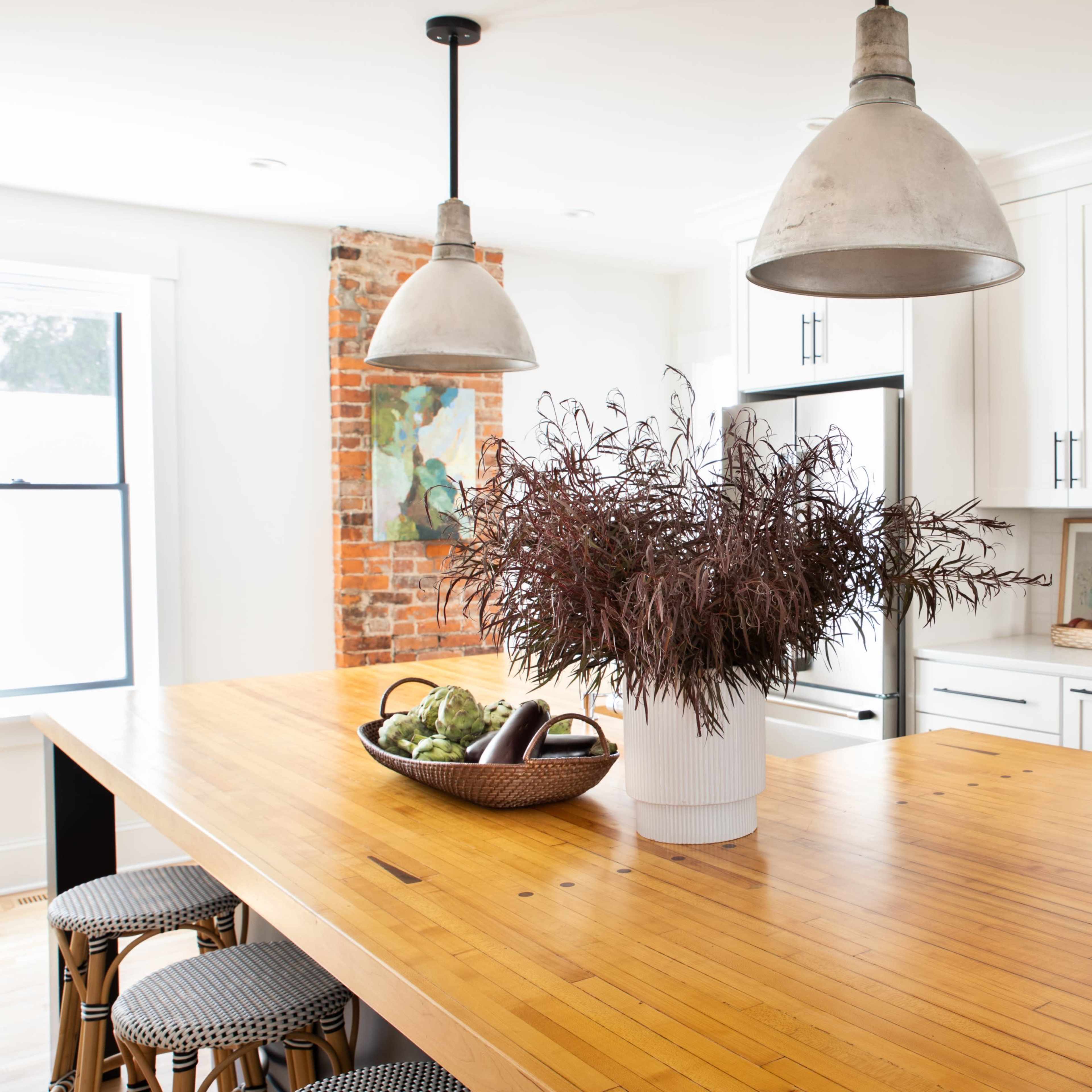 A modern kitchen features a large wooden island with a potted plant at the center, flanked by two pendant lights and surrounded by white cabinets and exposed brick walls.