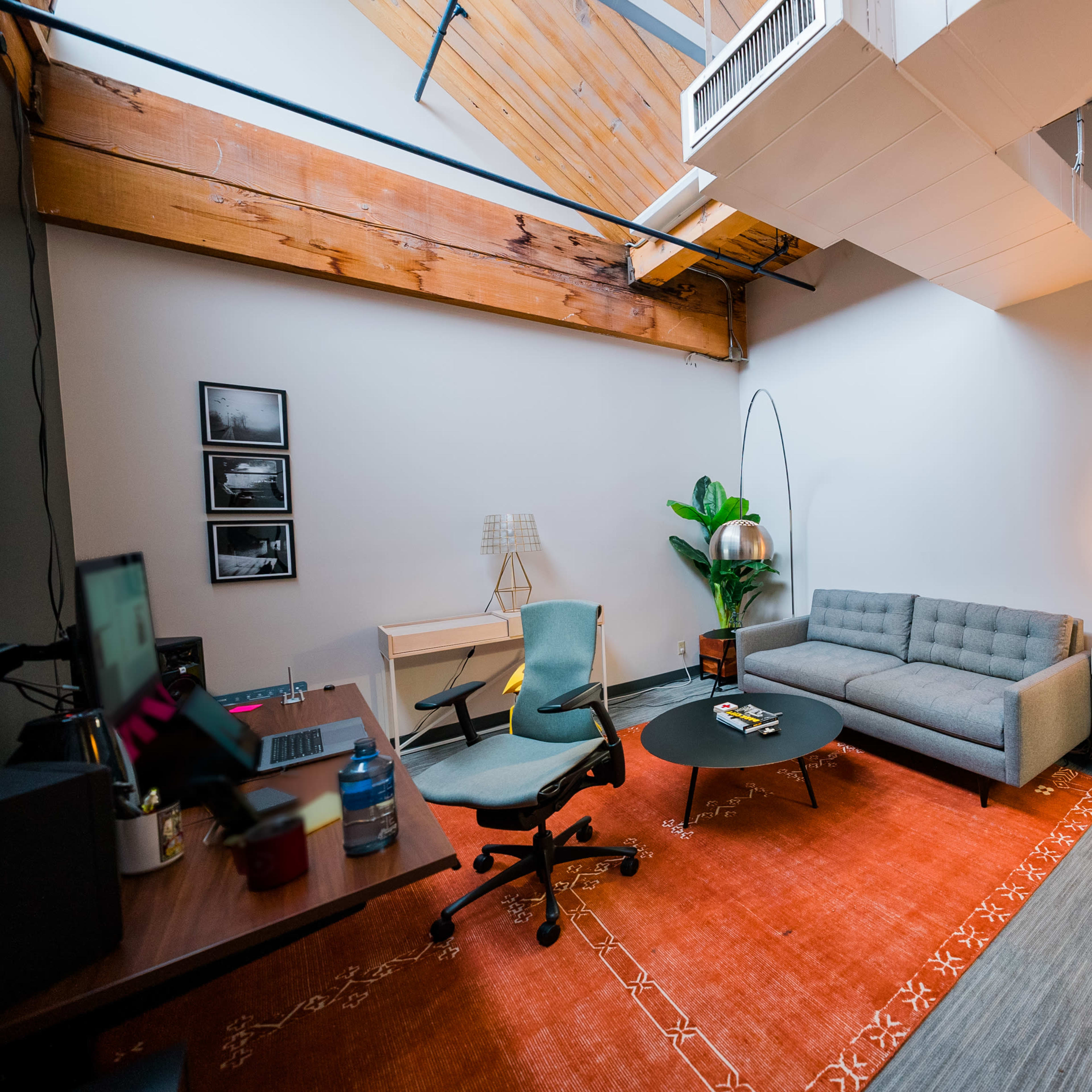 A modern office space features a gray sofa, a round black coffee table, a desk with an ergonomic chair, and various decorative elements including a plant and a warm light source.