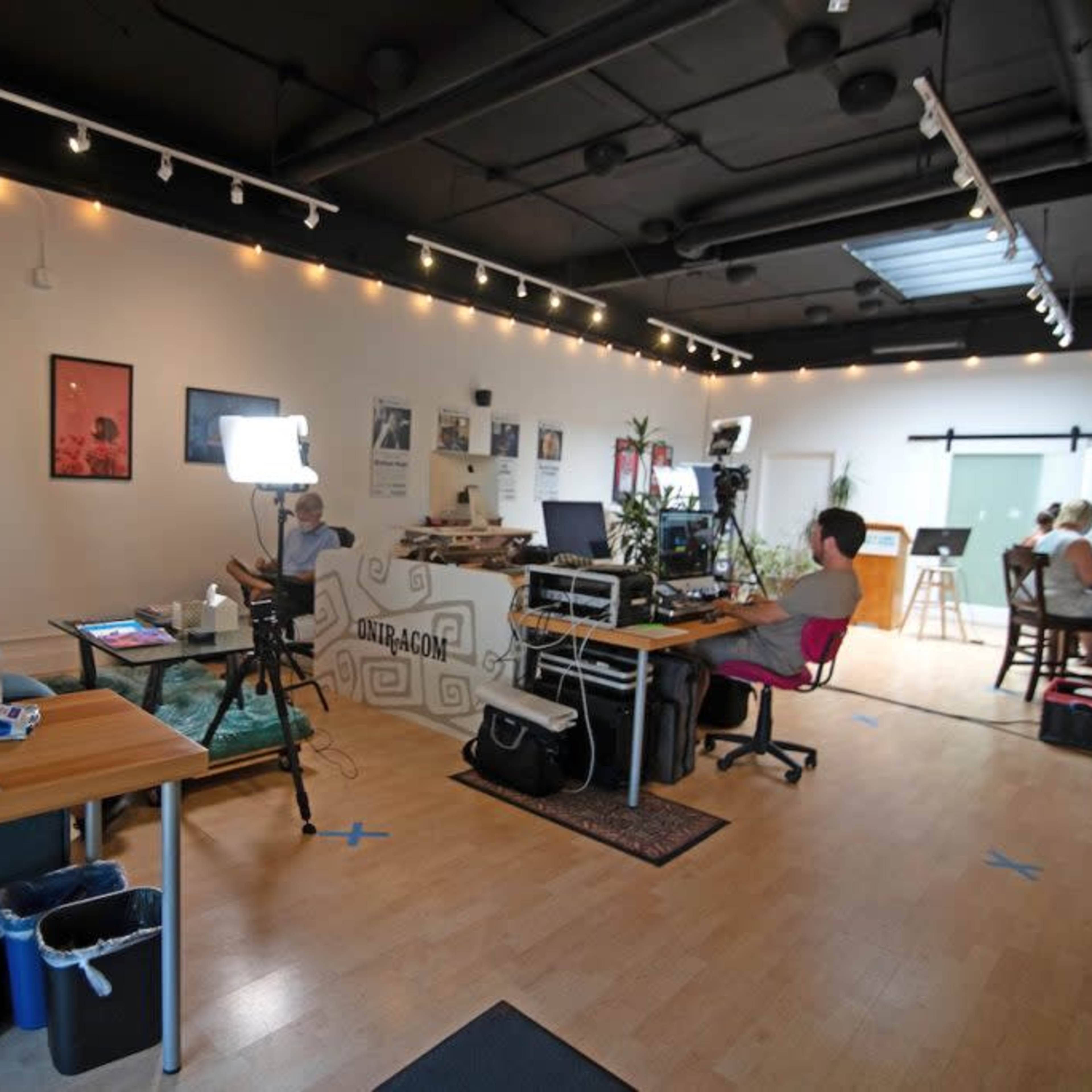 An open office space with multiple workstations, cameras set up for filming, and a few people engaged in various activities.