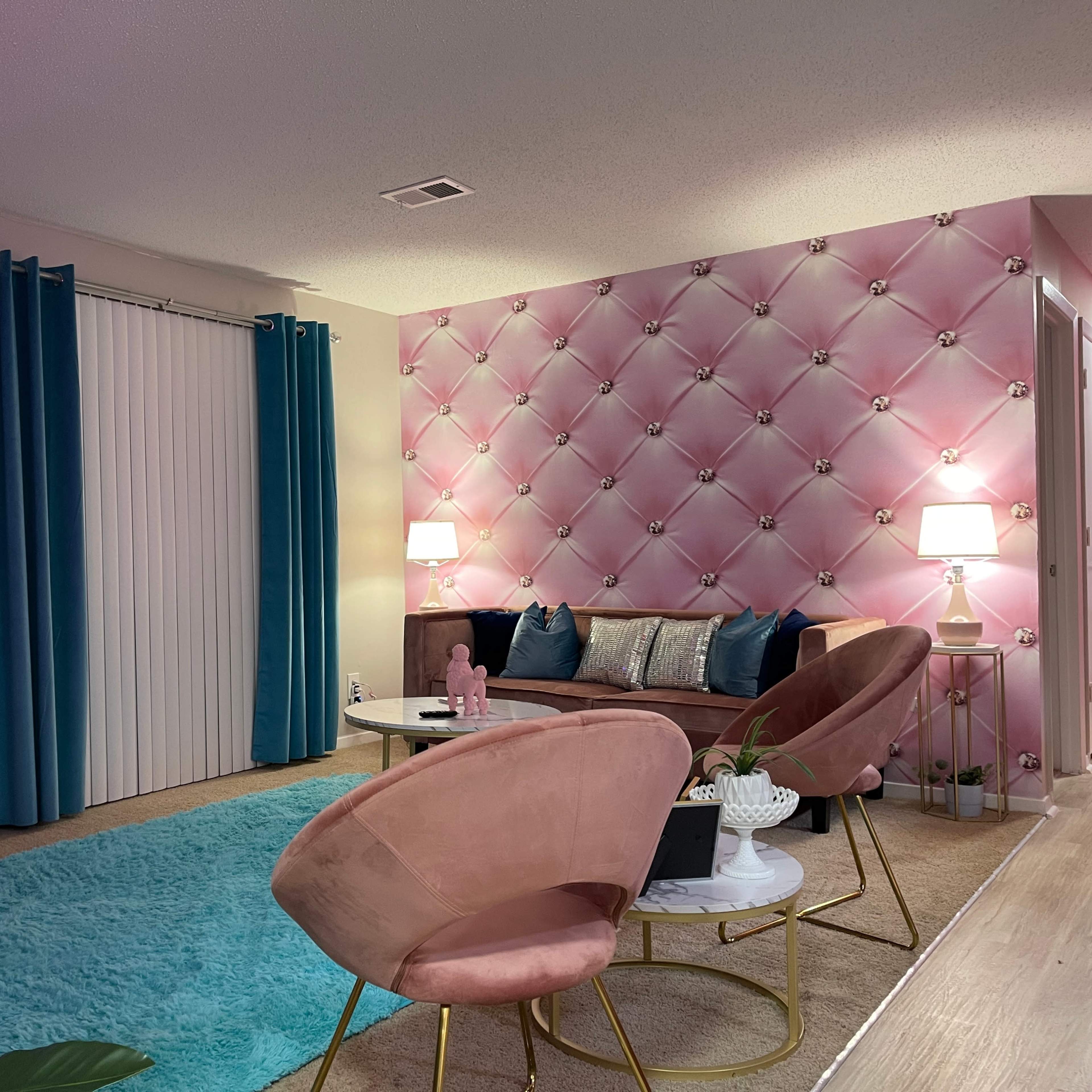 A modern living room featuring a pink tufted accent wall, teal curtains, a plush area rug, and stylish furniture including a sofa and two pink chairs.