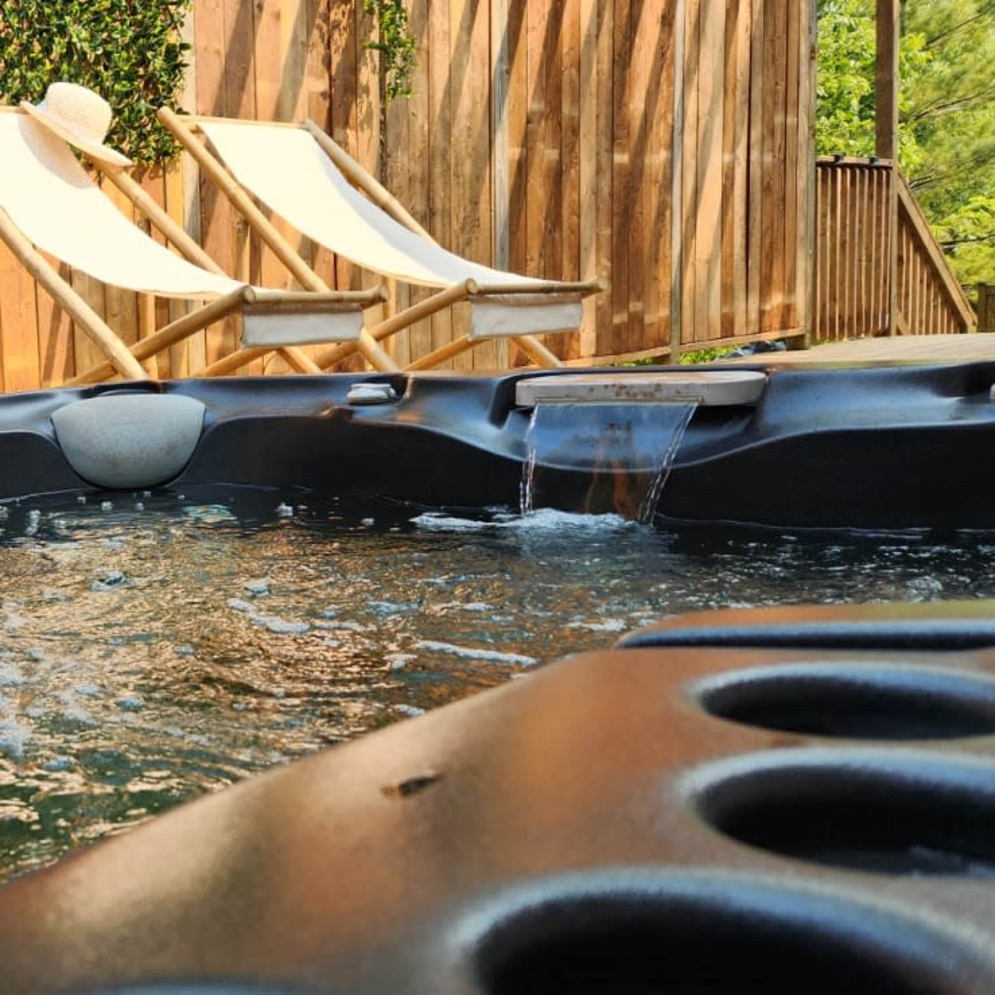 A hot tub with bubbling water next to two empty lounge chairs and a wooden fence covered in greenery.