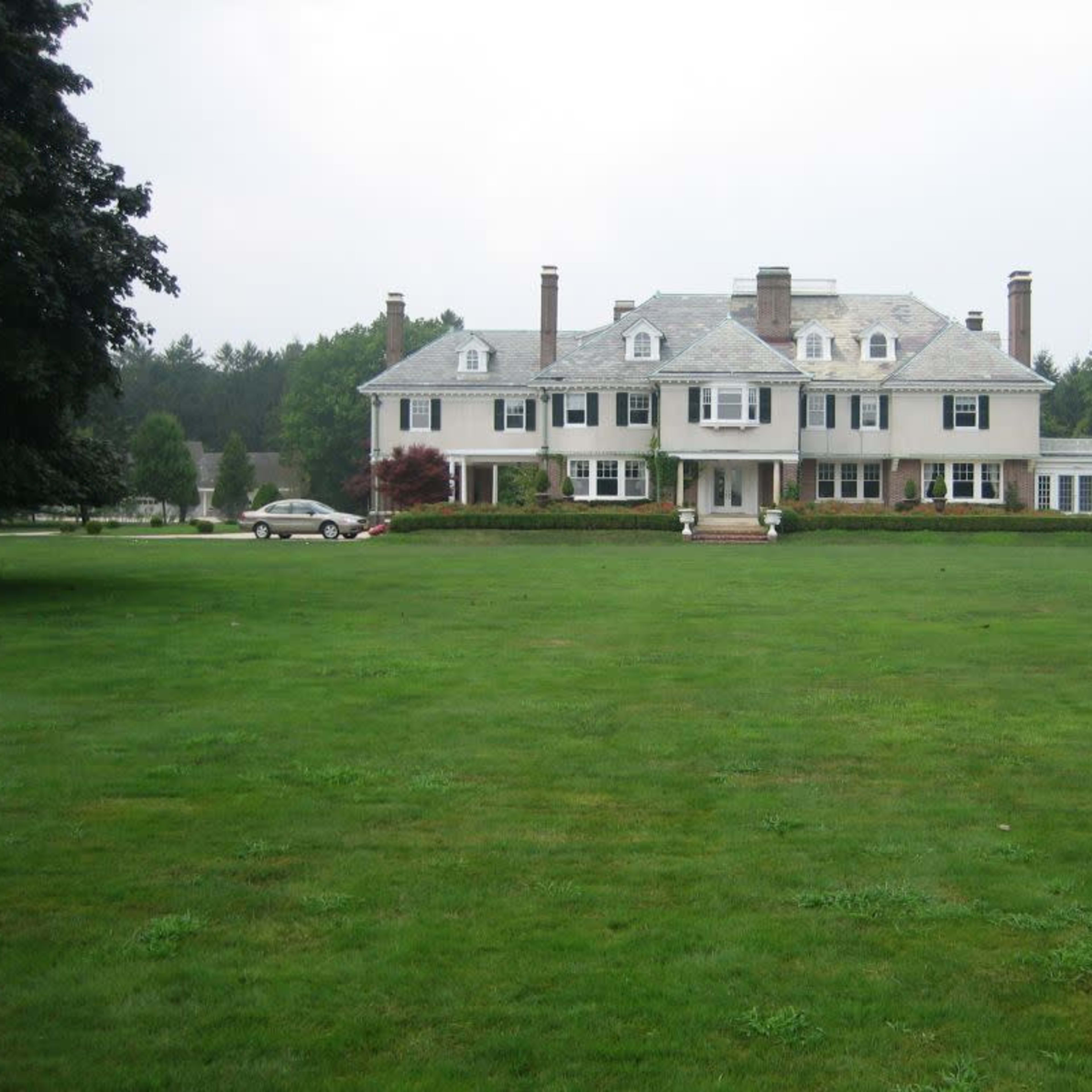 A large, two-story house with a gray roof and white exterior sits on a sprawling green lawn.