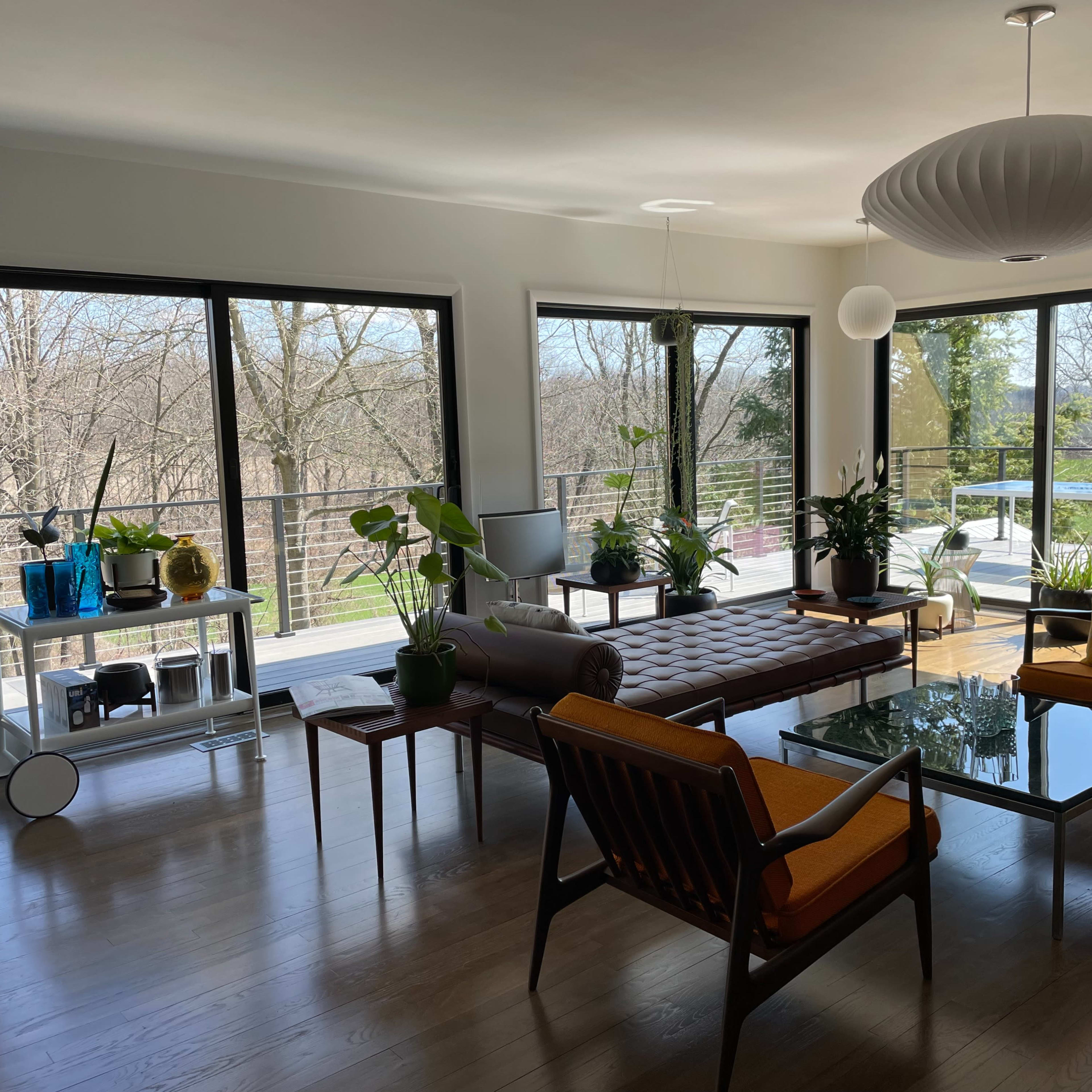 A modern living room with large windows overlooking a wooded area, furnished with a brown sofa, orange chairs, and various potted plants.