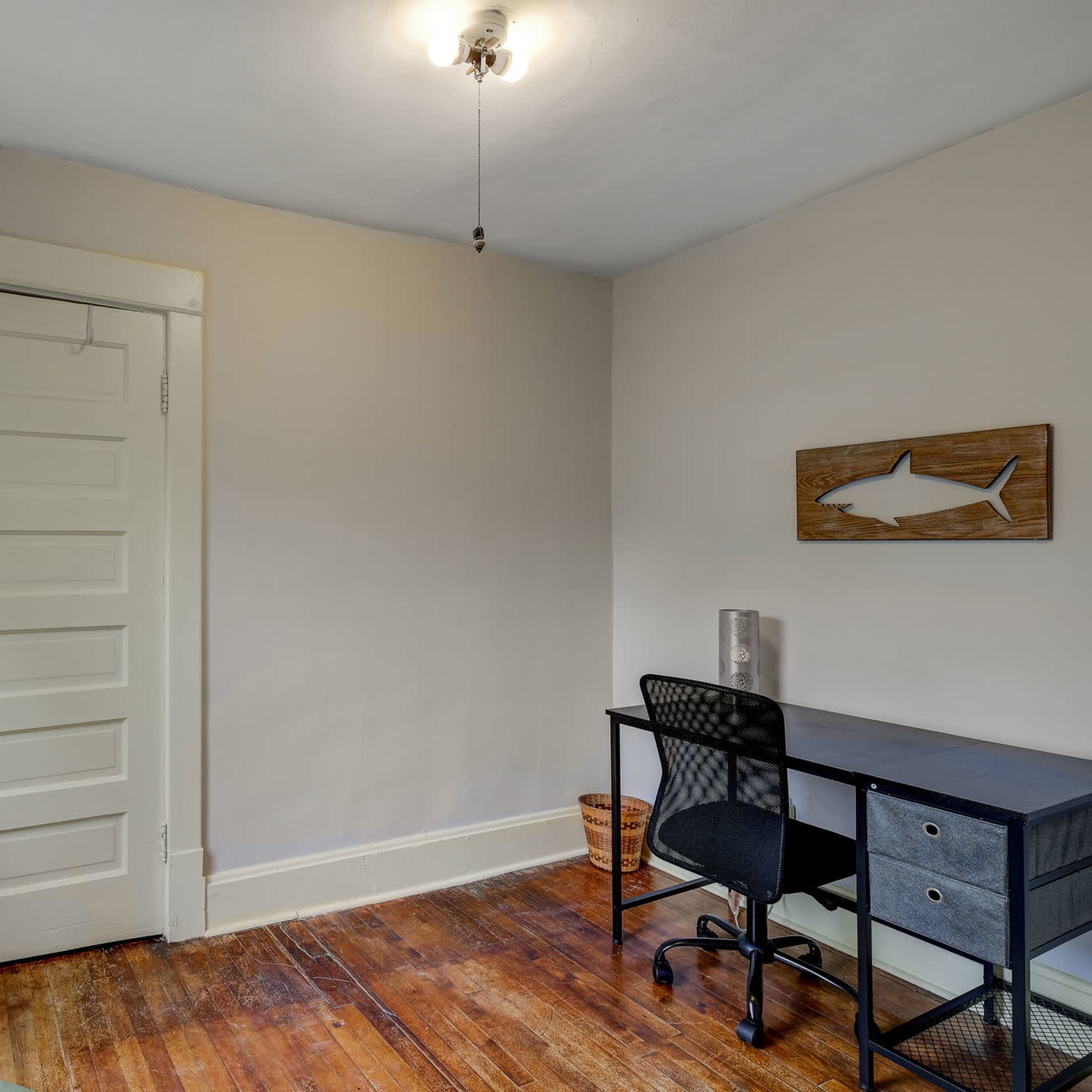 A simple, well-lit room featuring a desk, a chair, a wooden door, and a small wall decor item resembling a fish.