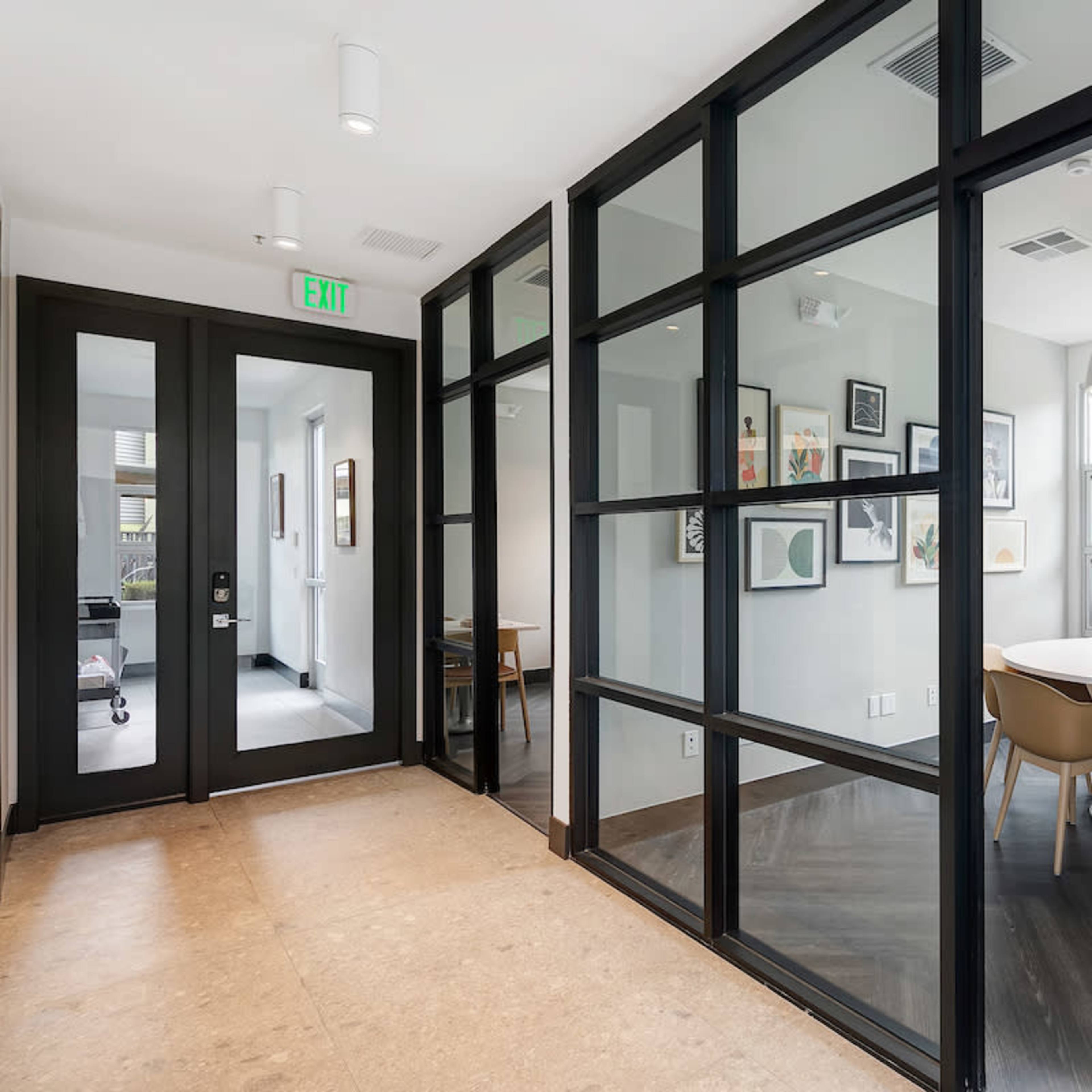 A hallway features black-framed glass walls that lead to a modern meeting room with a round table and chairs.