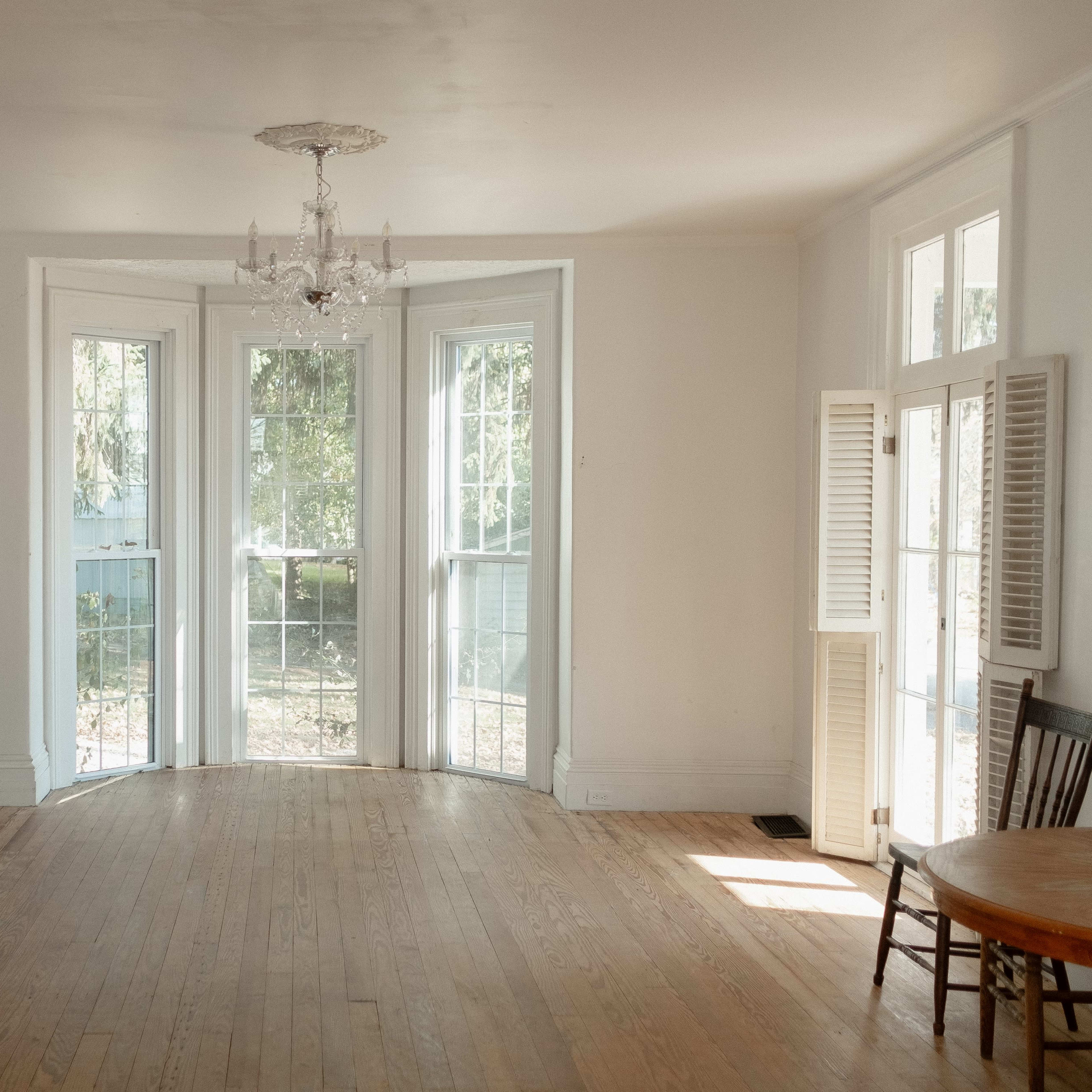 A bright, empty room features large windows, wooden floors, a chandelier, and a decorative fireplace.
