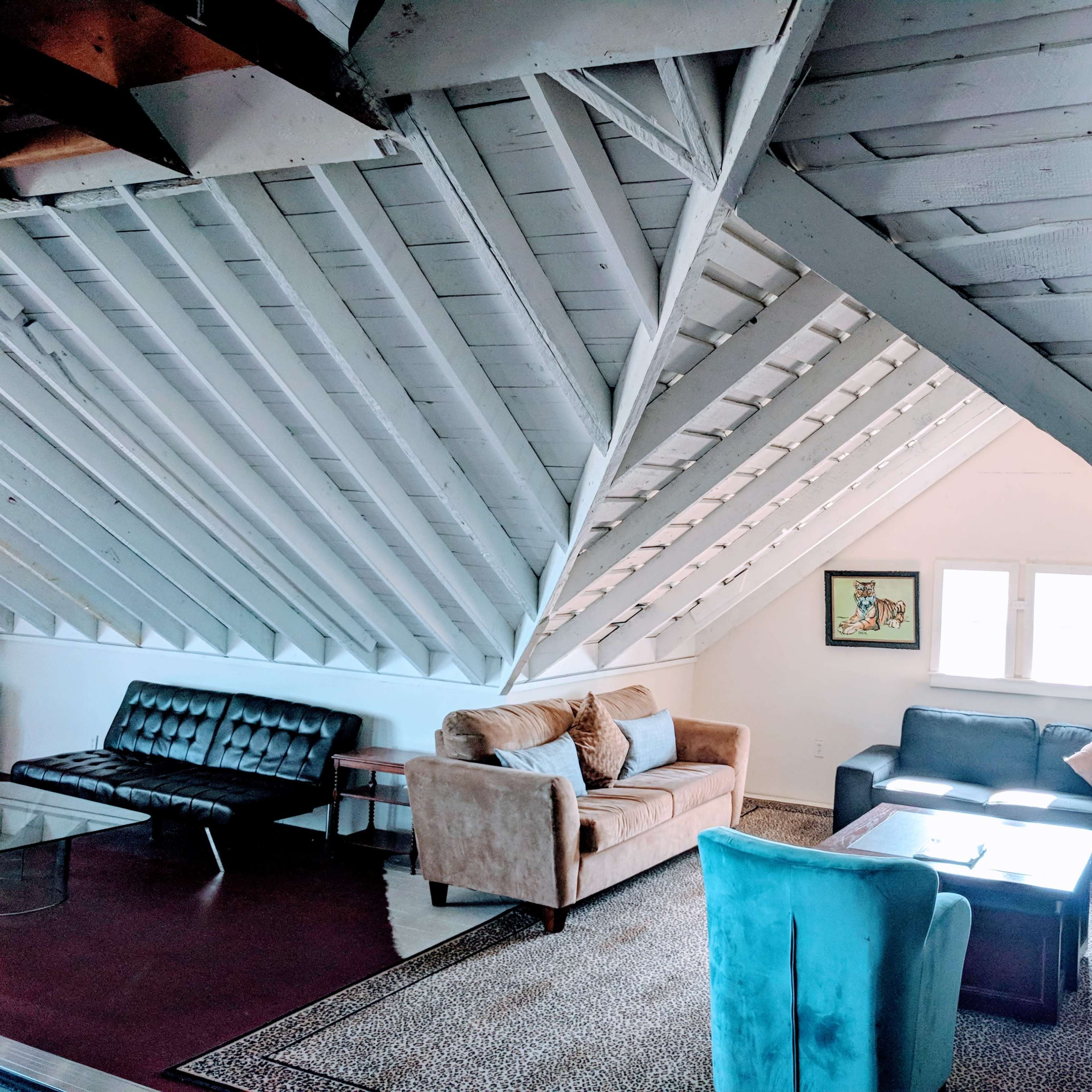A loft-style living room with slanted wooden ceilings, showcasing a mix of seating options including a brown sofa, gray chairs, and a black futon arranged around a central table.