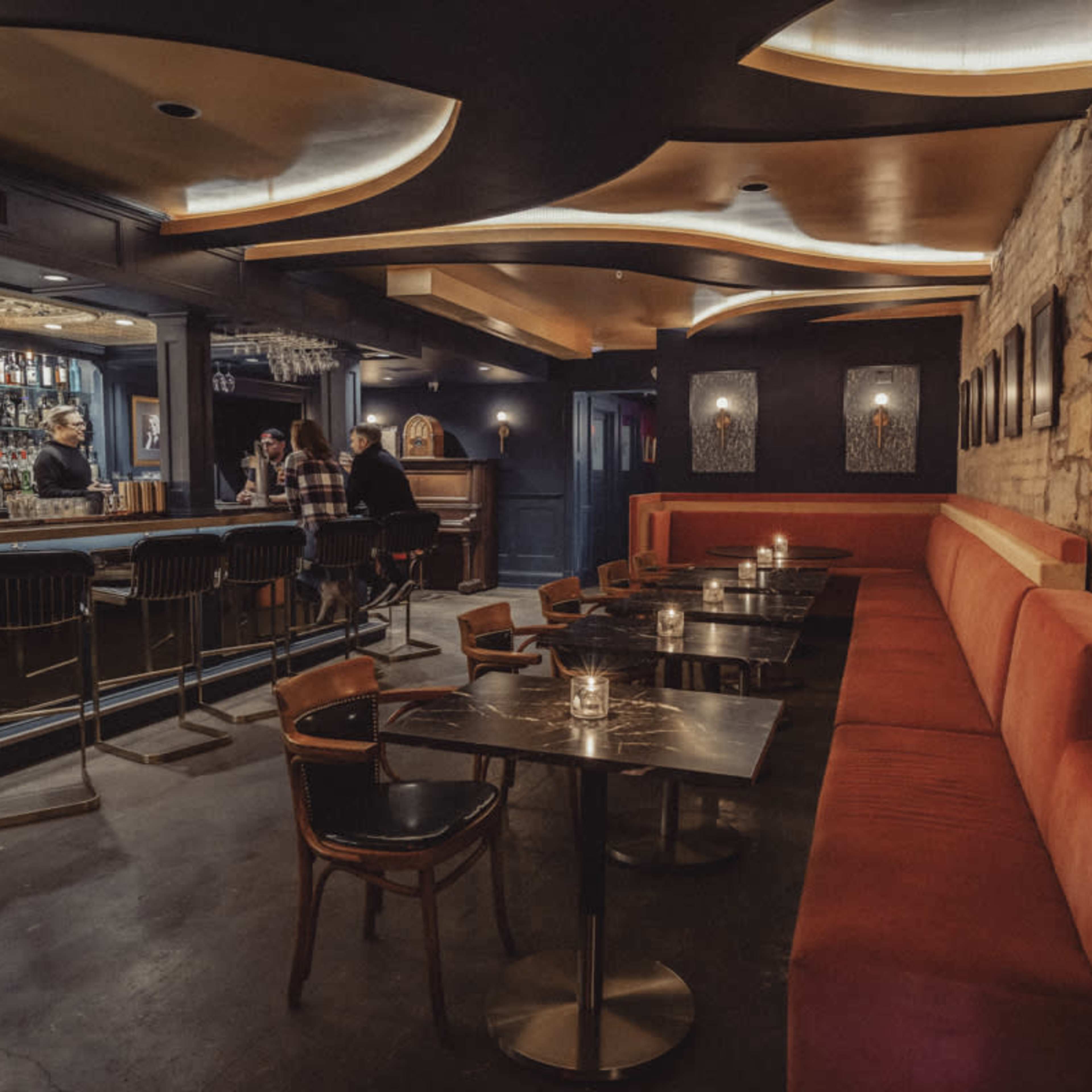 A dimly lit bar interior featuring a curved ceiling, a stone wall, and a seating area with red velvet booths and small tables.