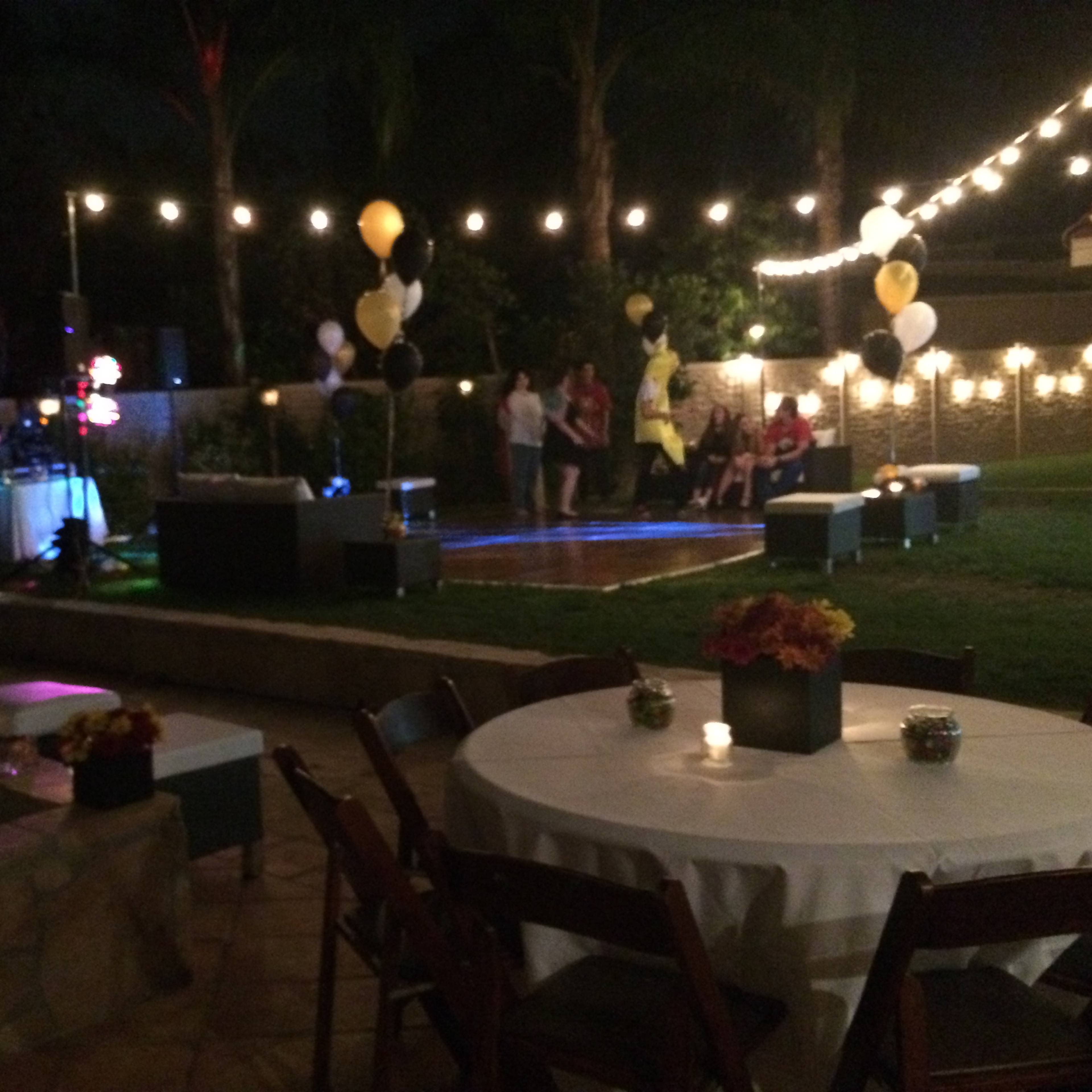 A nighttime outdoor party atmosphere with tables, string lights above, a small dance area, and guests mingling among decorations.