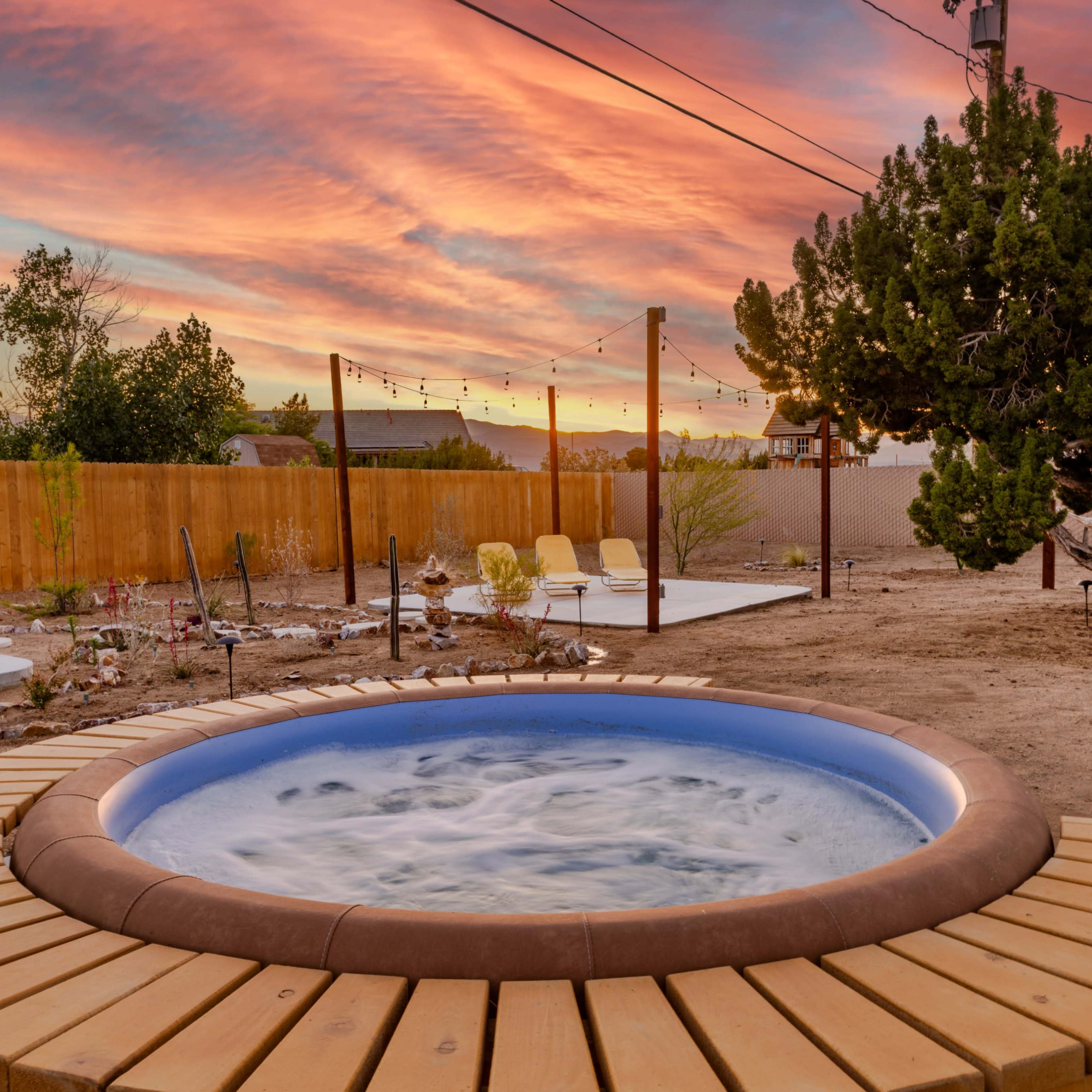 A hot tub with a wooden deck sits in a backyard at sunset, surrounded by trees and a wooden fence.