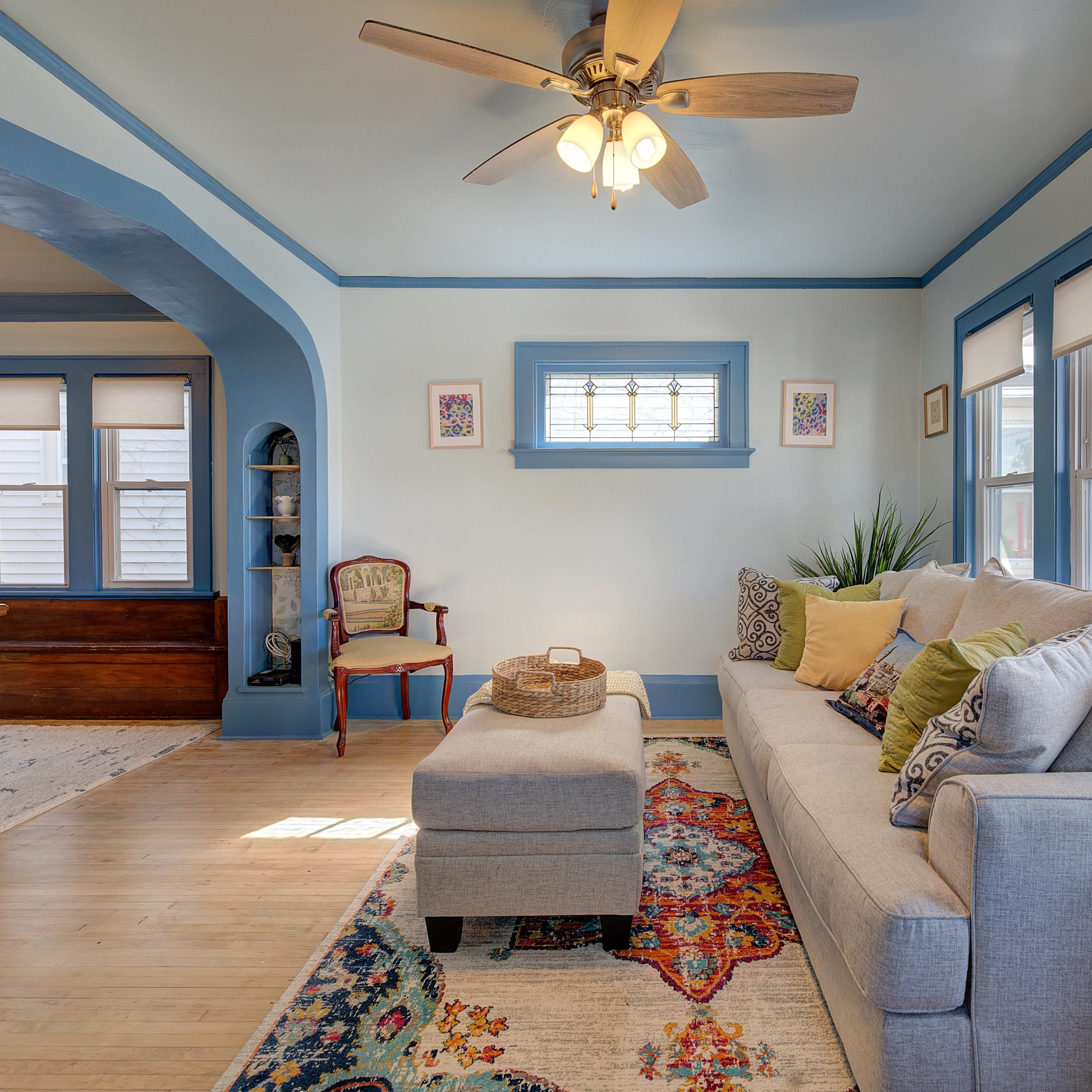 A living room with a light blue color scheme, featuring a beige sofa, an ottoman, decorative pillows, and large windows that allow natural light to illuminate the space.