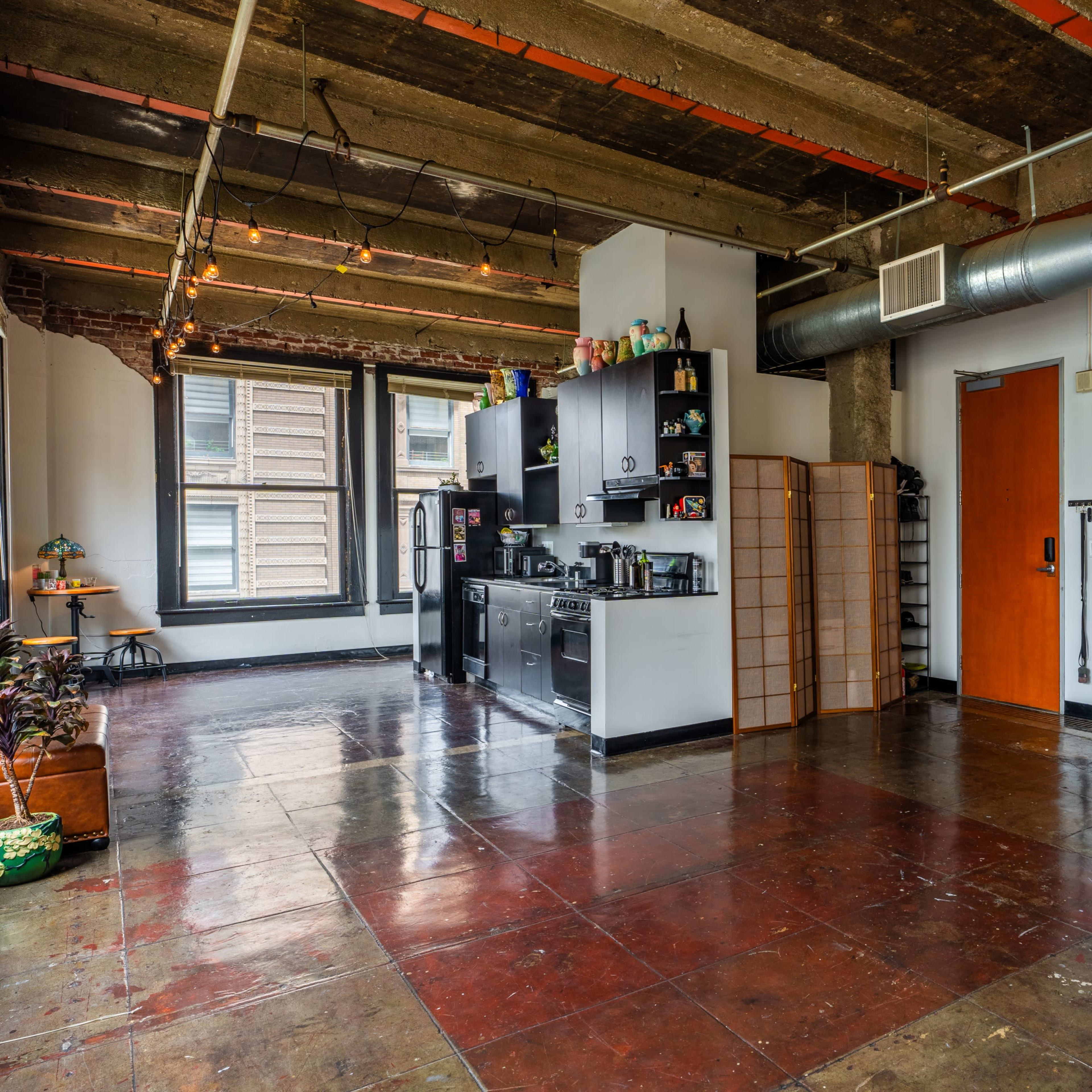 An open loft space features a kitchenette along one wall, a wooden door leading to another area, and various decorative elements, including a lamp and plants.