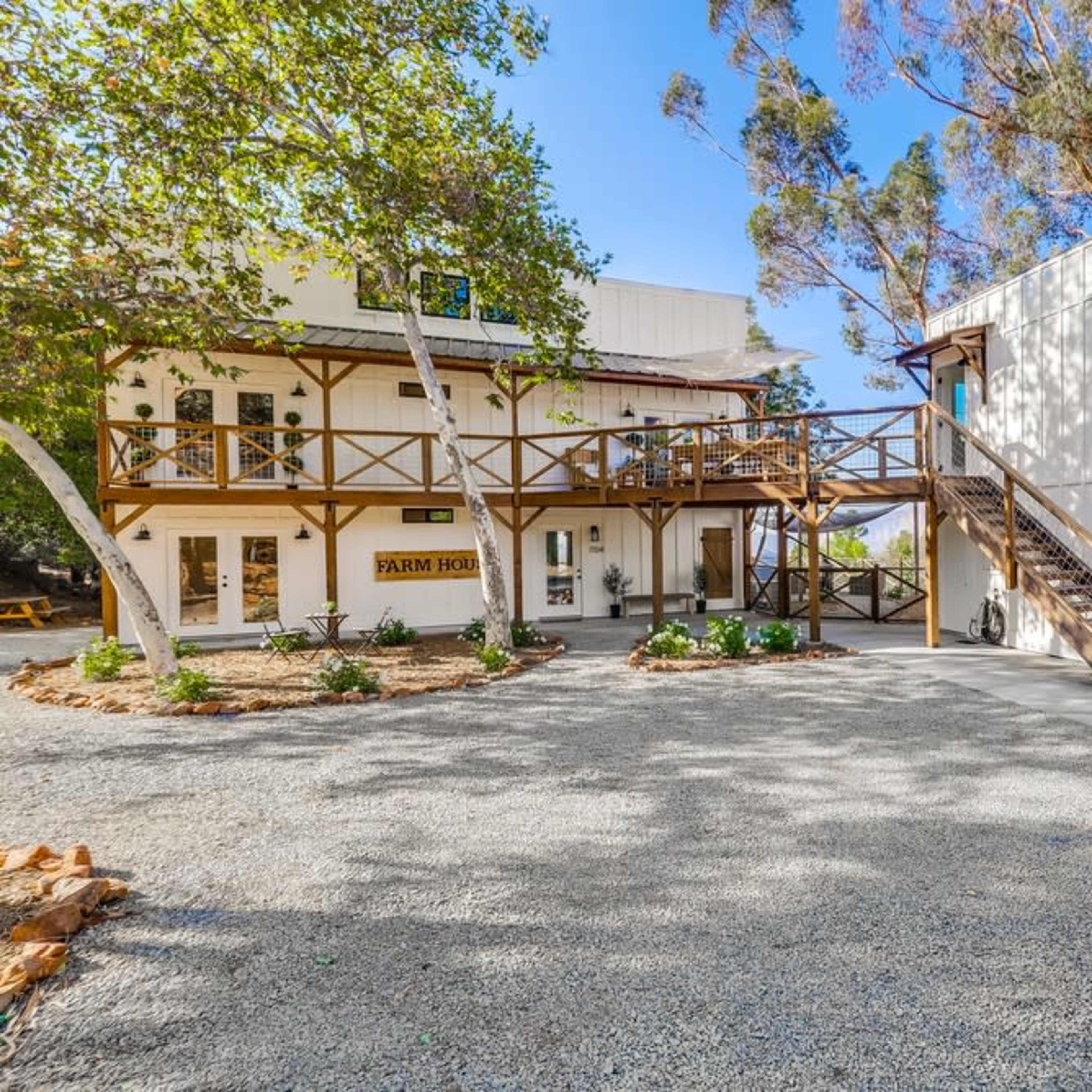 A two-story farmhouse with a wooden deck and stairs, surrounded by gravel and greenery.