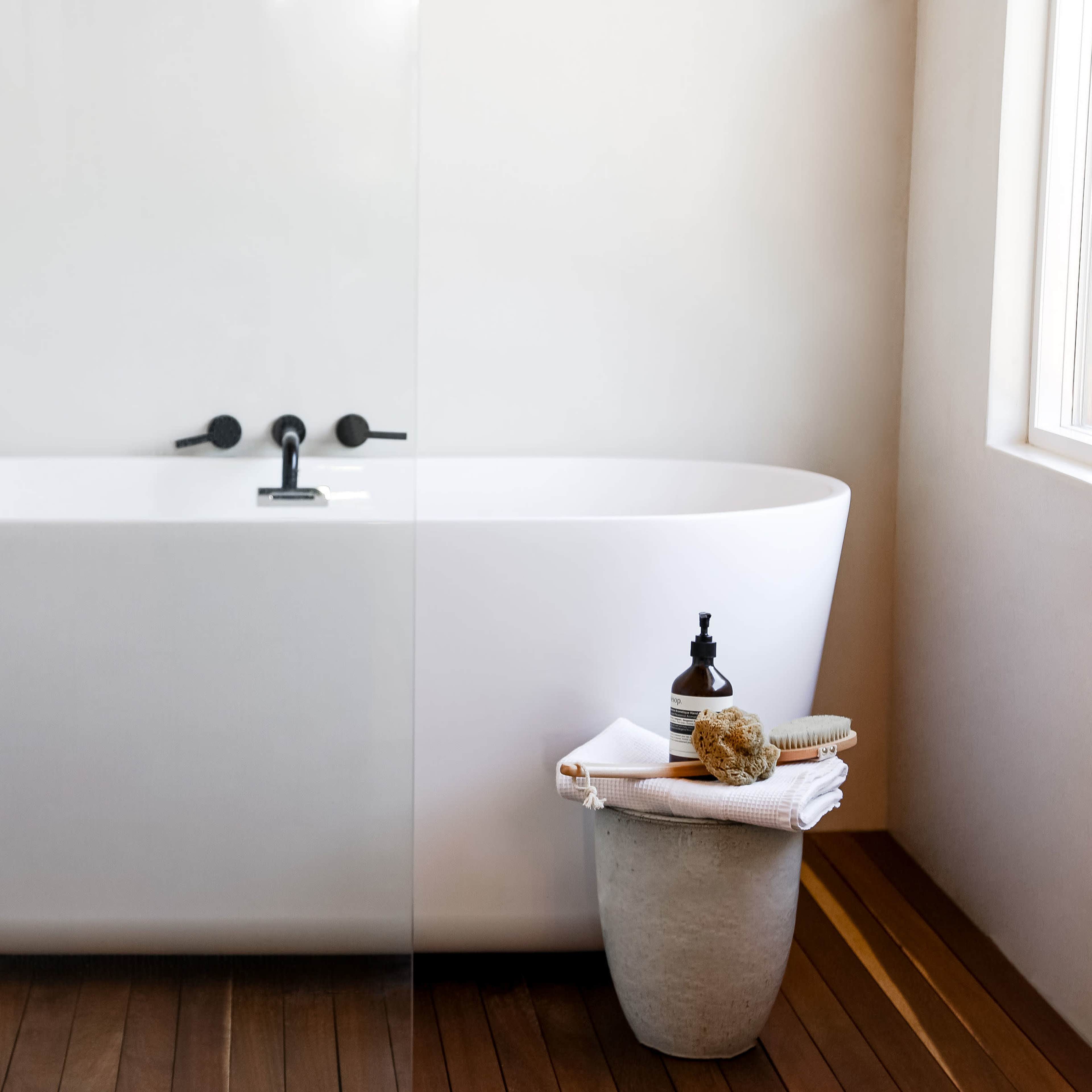 A modern bathroom features a white freestanding bathtub next to a stool holding towels, a sponge, and a bottle.