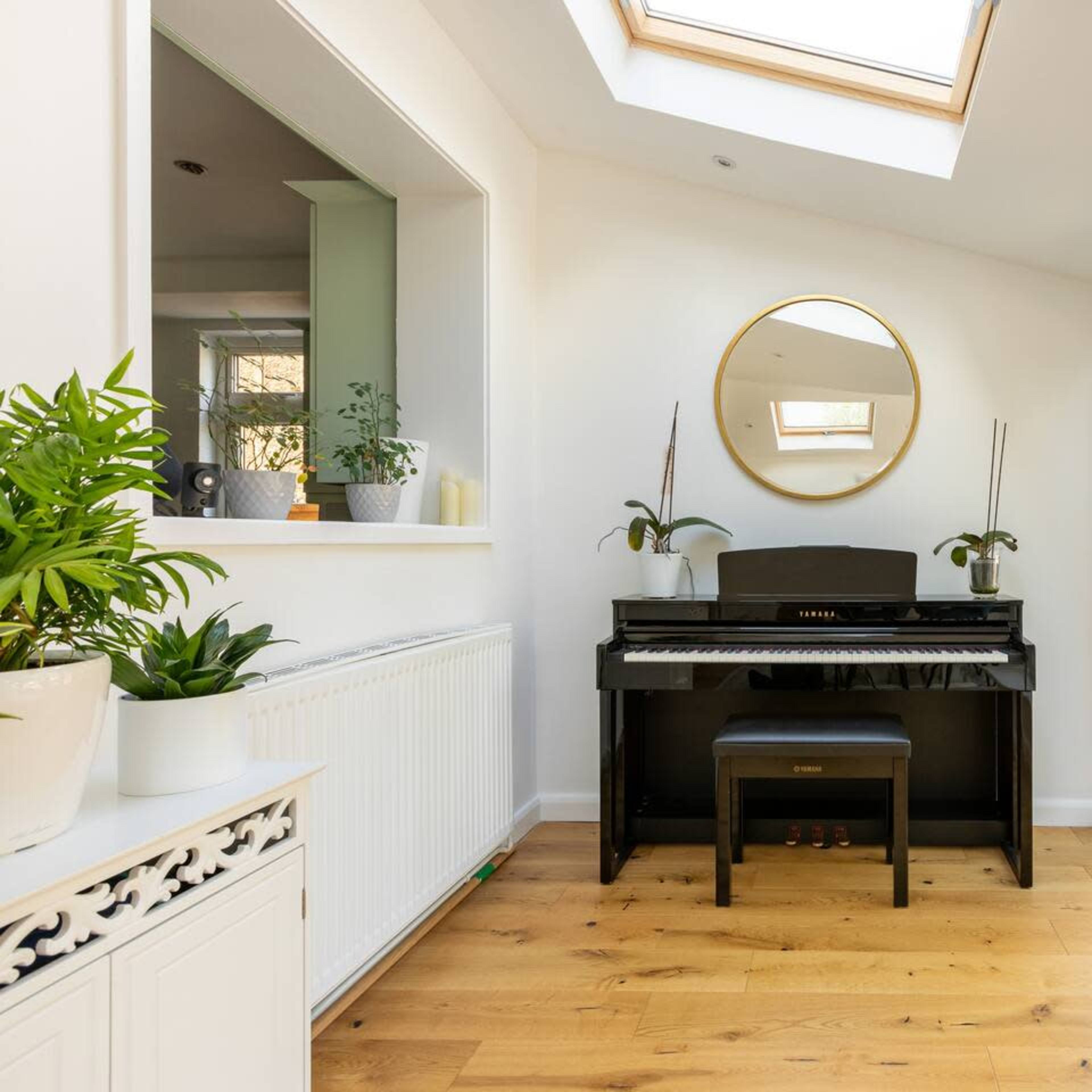 A black piano sits against a wall under a skylight, surrounded by large windows and potted plants in a bright, airy room.