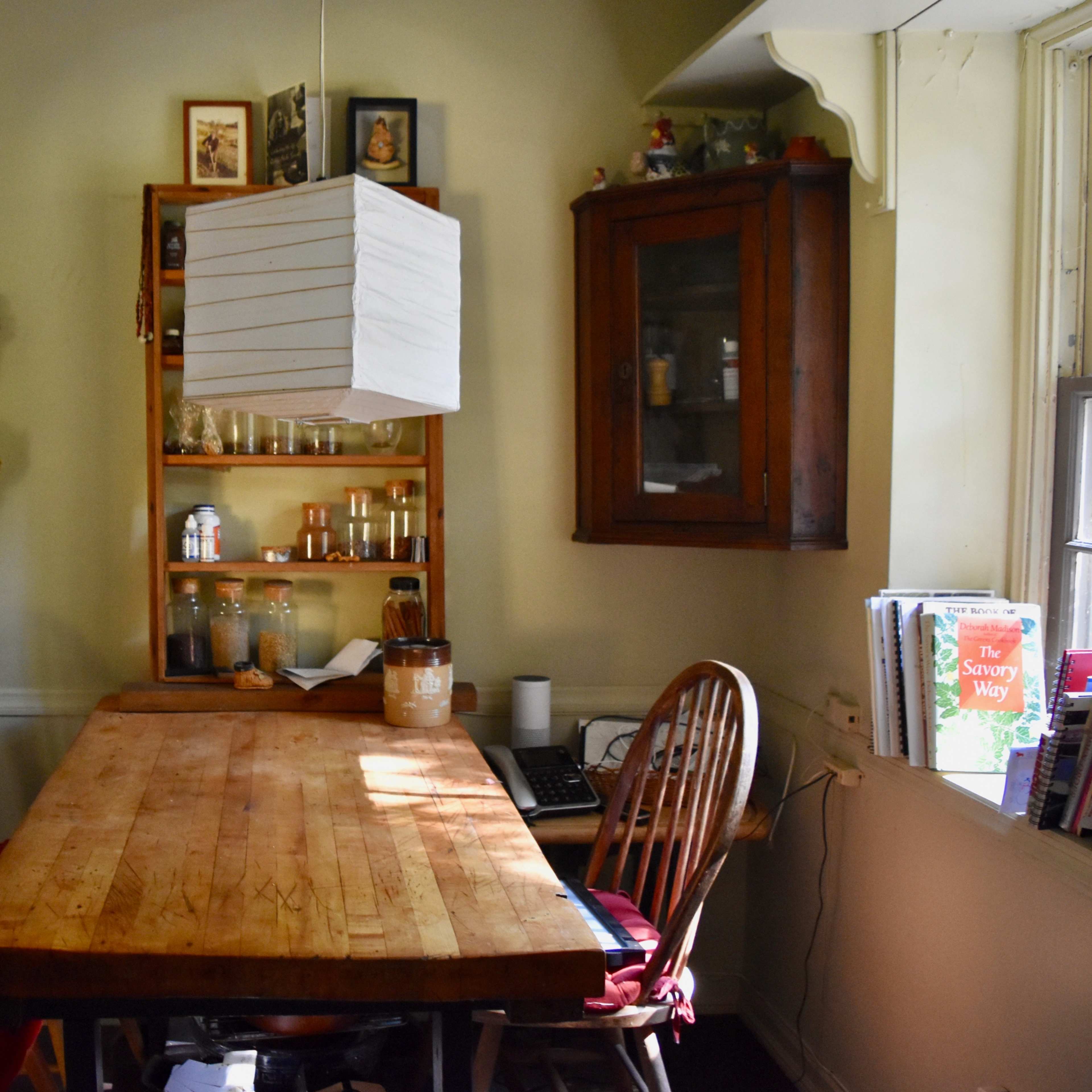 A wooden table sits in a cozy dining area surrounded by chairs, a bookshelf, and a cabinet with glass jars.