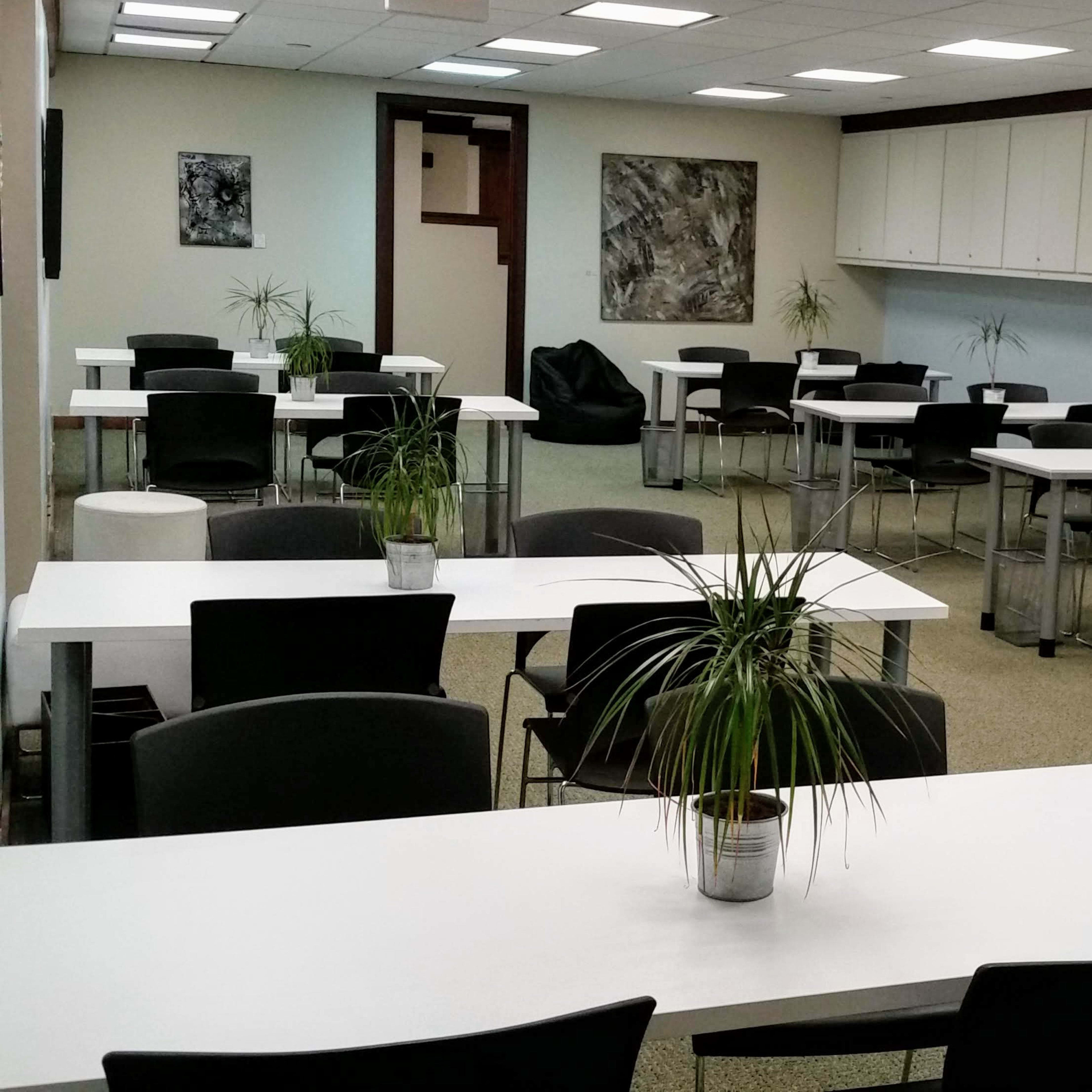 A spacious classroom or meeting room with several white tables and black chairs, featuring potted plants on each table and a neutral-colored carpet.