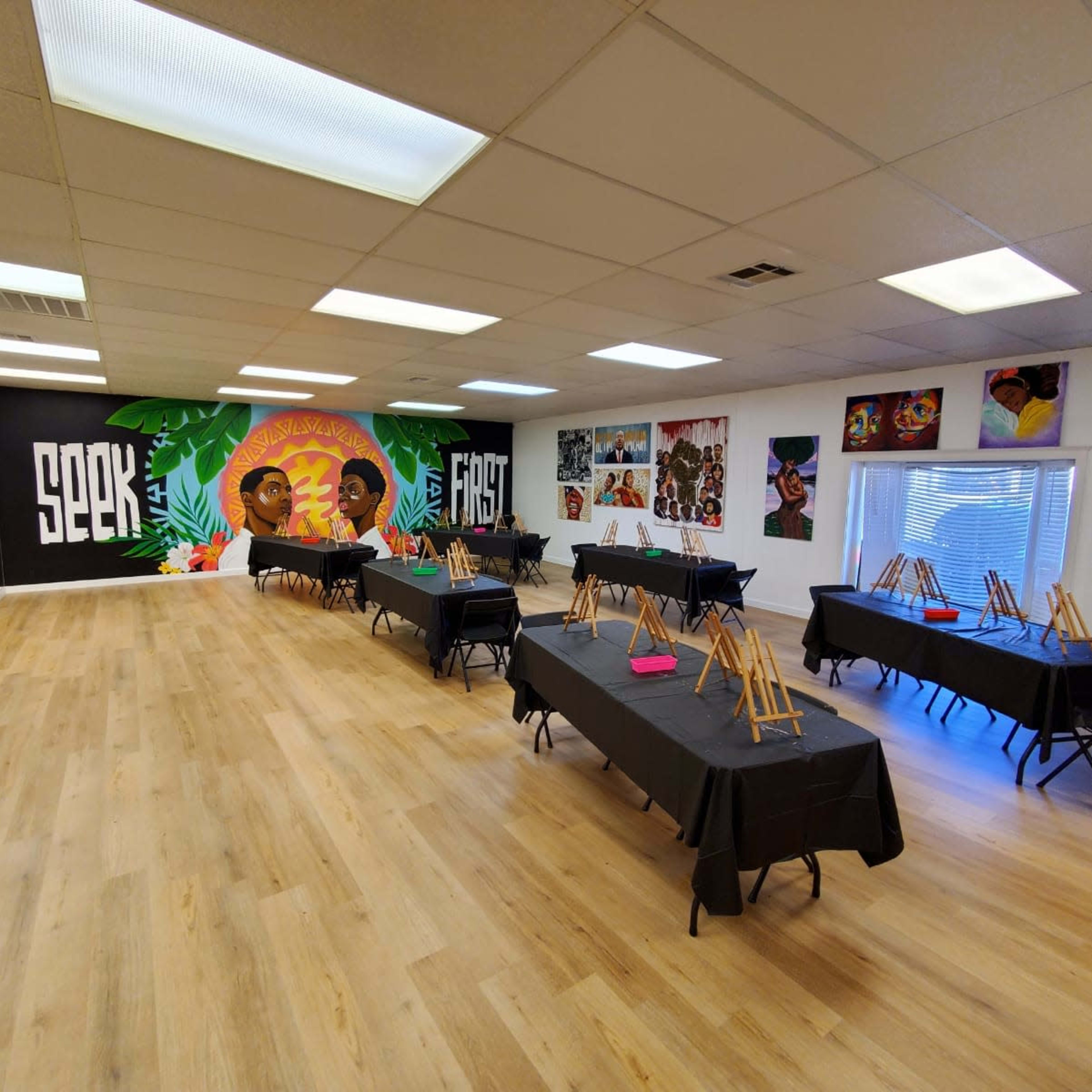 An art studio with several black tables set up for an event, each equipped with easels, surrounded by colorful murals and artwork on the walls.