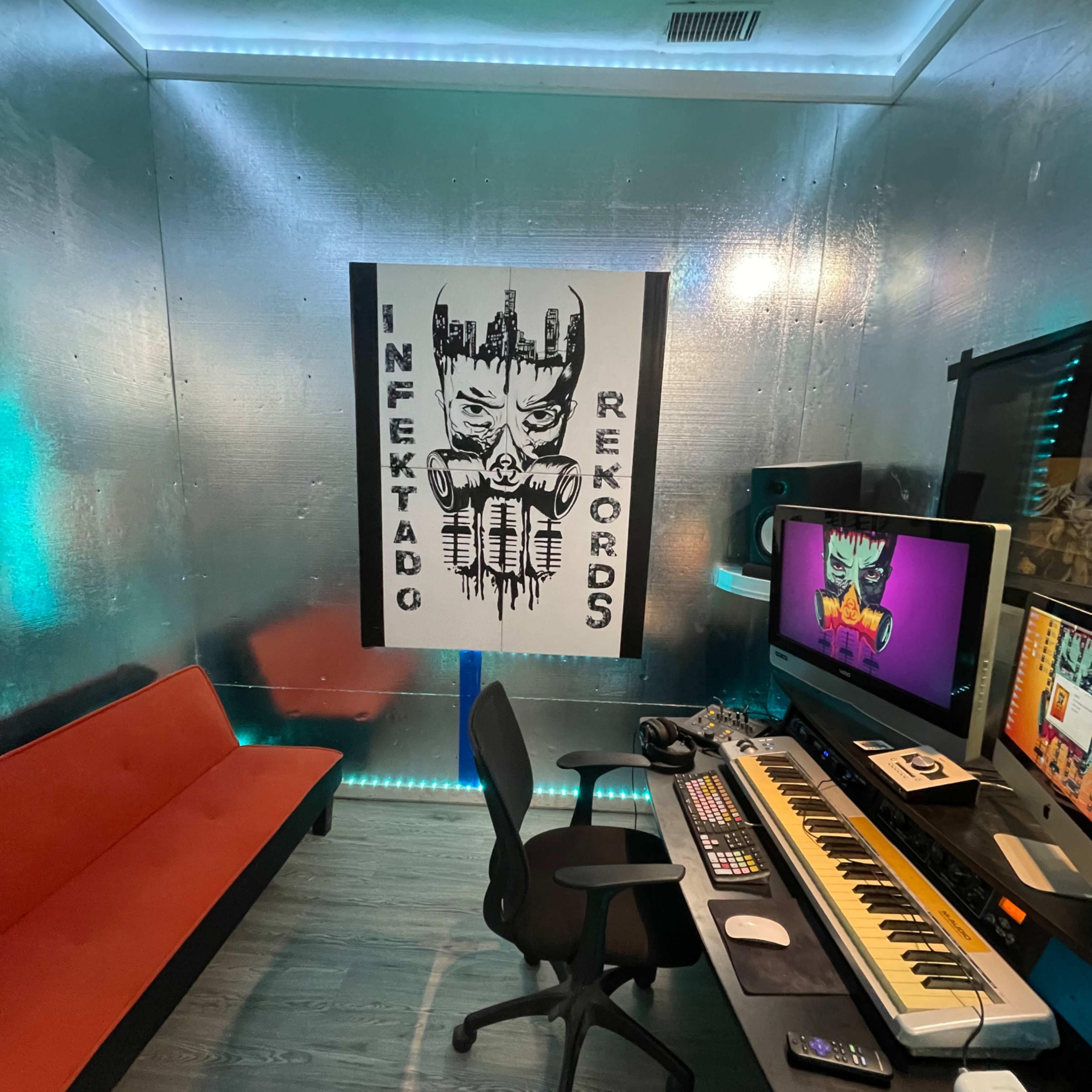 A small music studio with a red couch, a keyboard, and a computer setup against a silver metallic wall featuring a graphic poster.