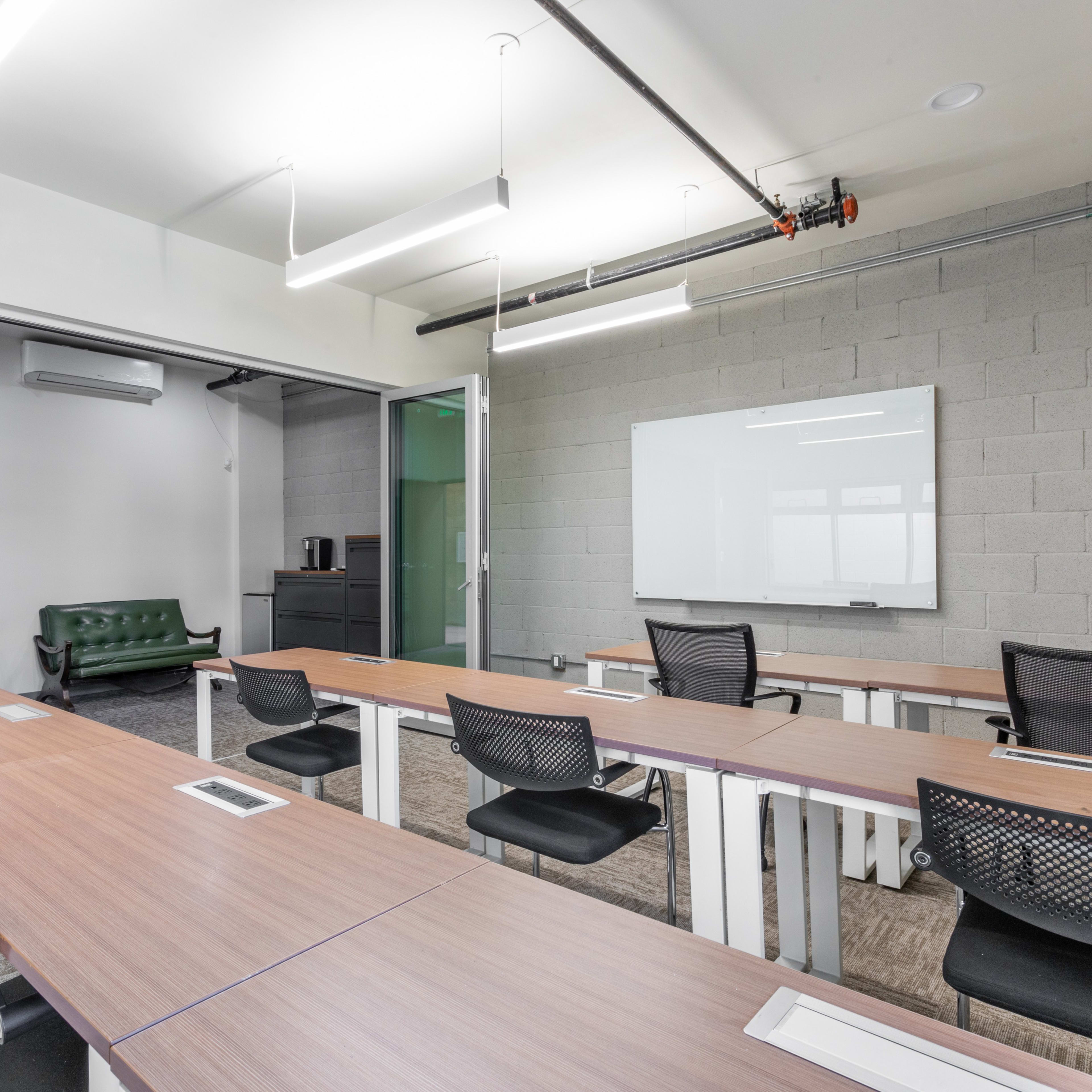A modern classroom with multiple tables, black chairs, a green couch in an adjoining room, and a whiteboard on the wall.