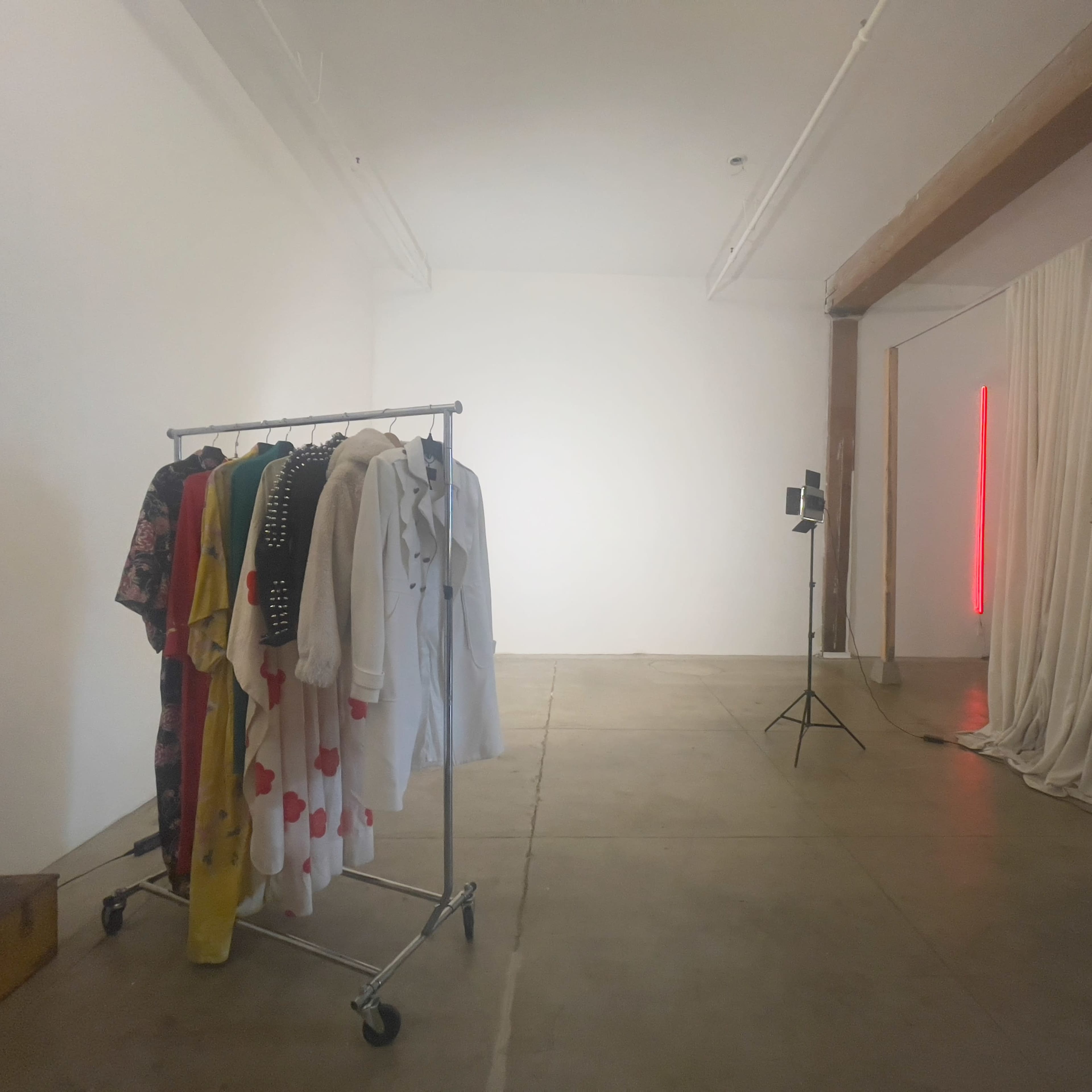 A spacious room with a clothing rack holding various garments, a light stand, and a curtain draped along one wall.