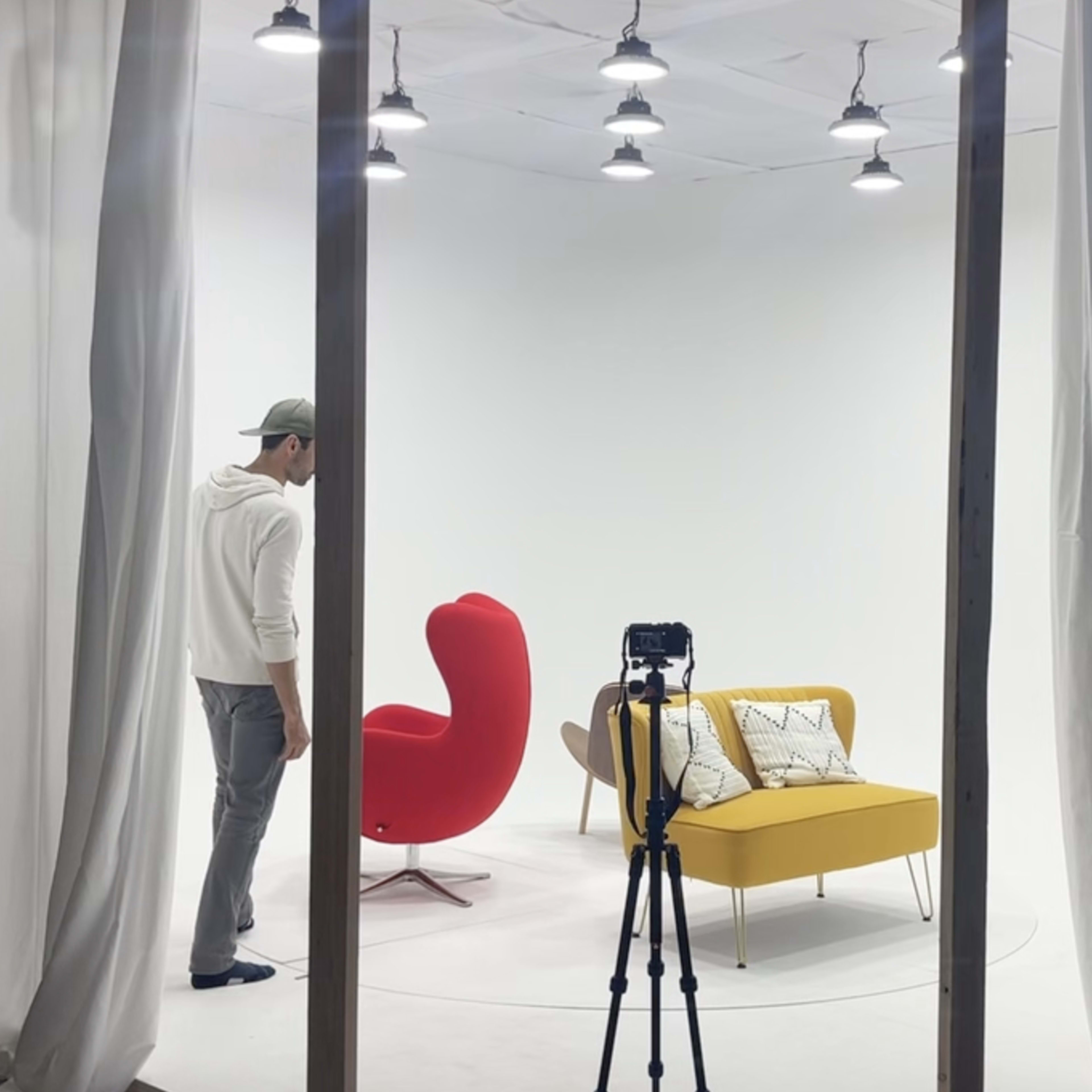 A person stands in a minimalistic studio with two brightly colored chairs and a camera on a tripod.