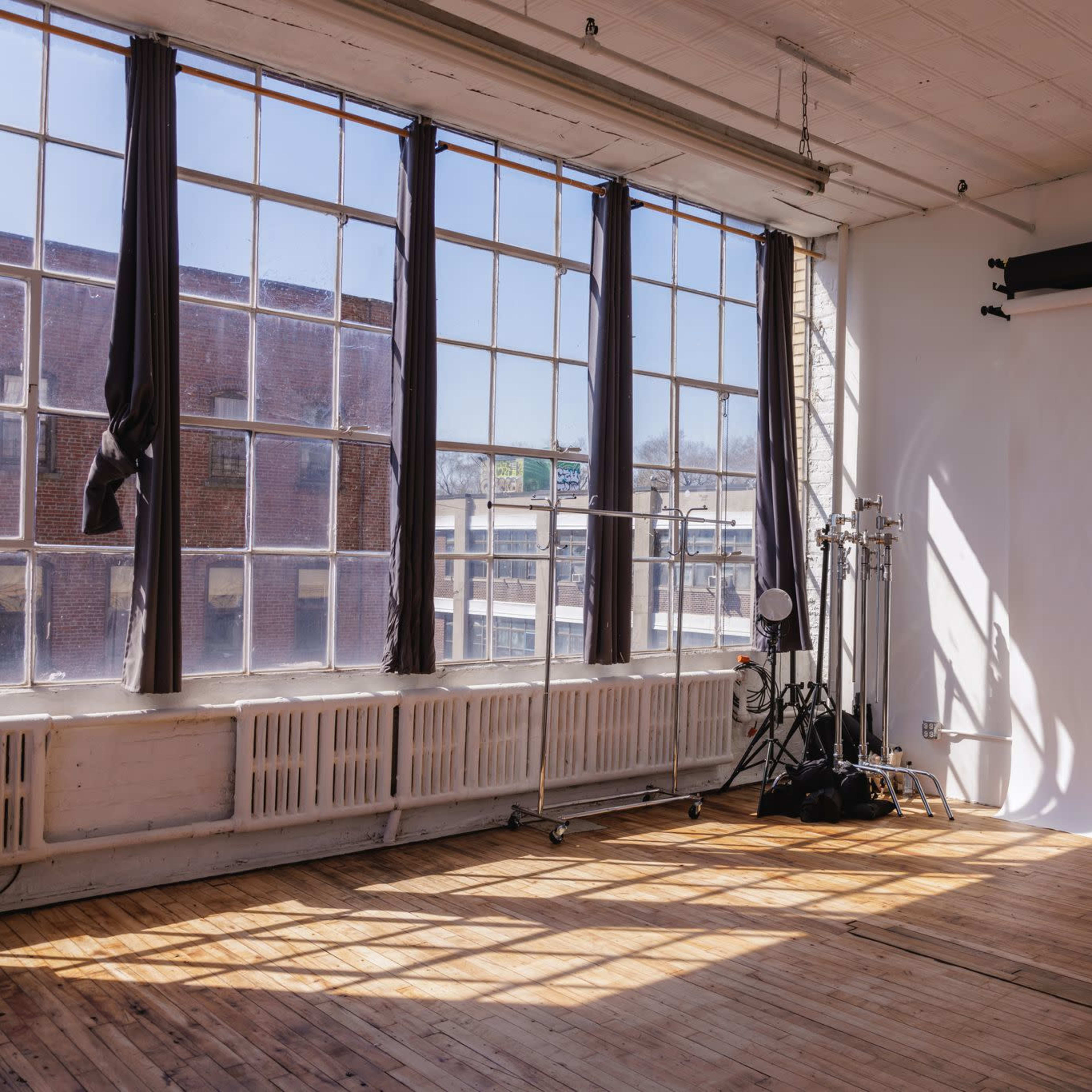 A bright photography studio features large windows, wooden flooring, and a white backdrop with lighting equipment positioned nearby.