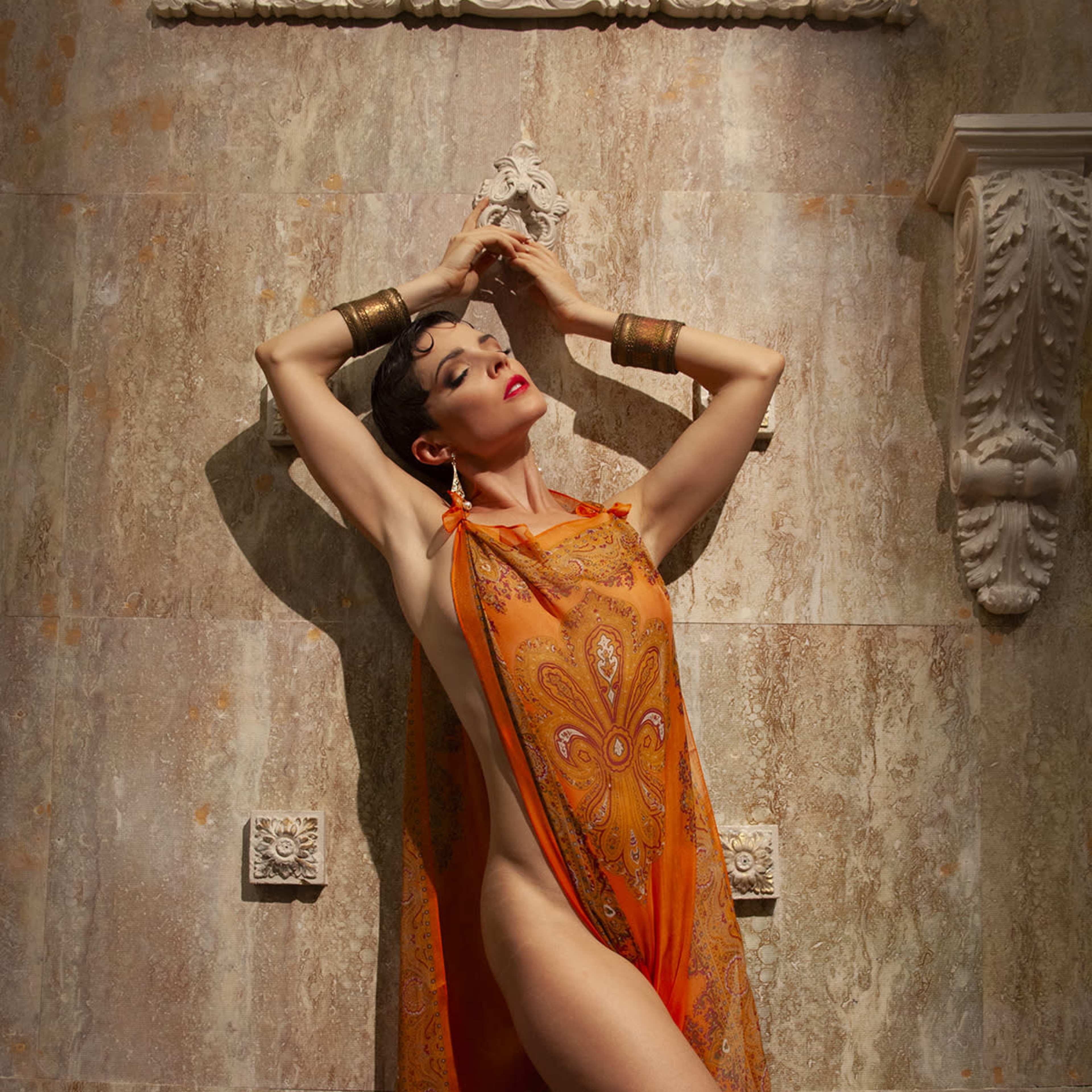 A model poses against a decorative wall while wearing an orange patterned gown that drapes elegantly.