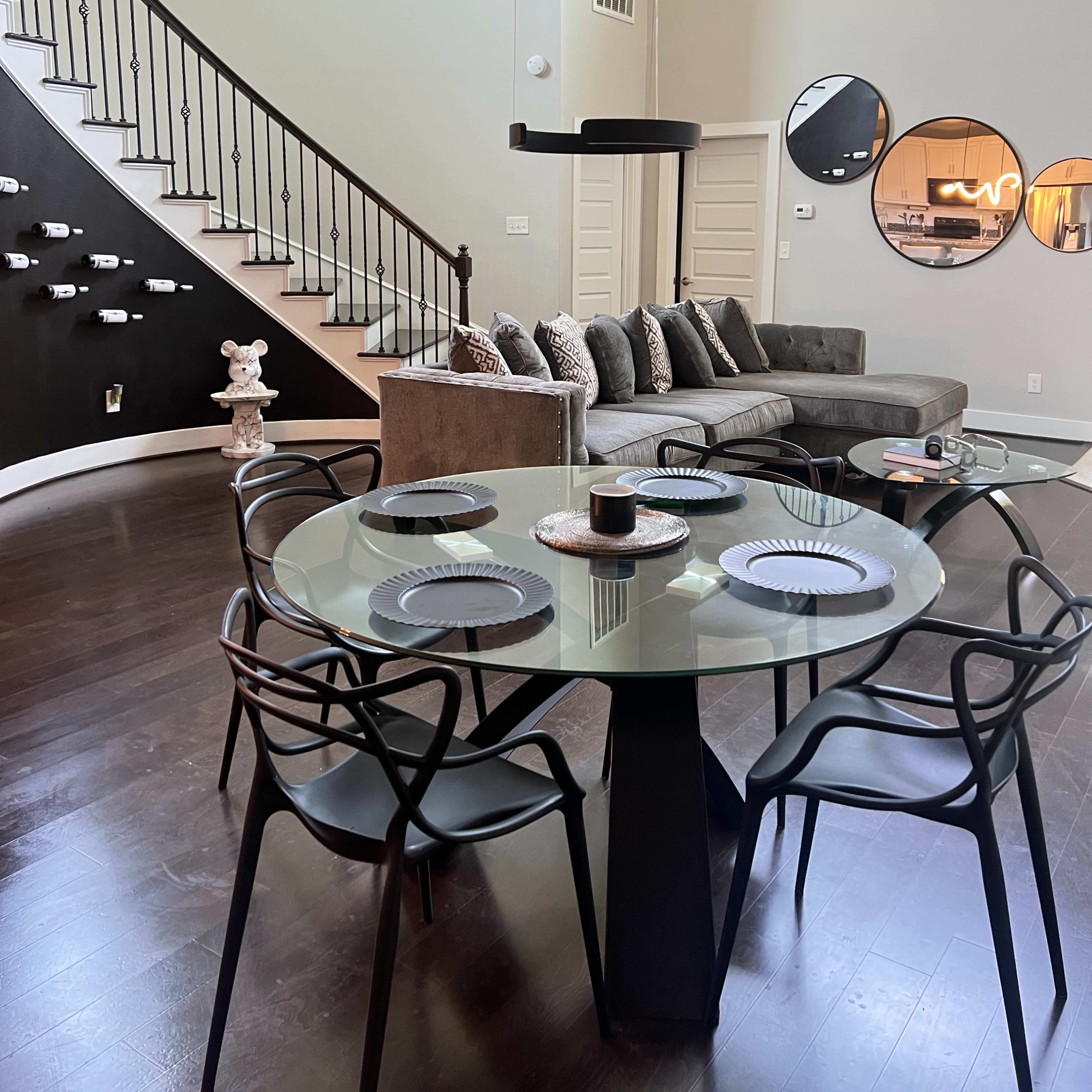 A modern living space features a dining area with a glass table and black chairs adjacent to a curved staircase leading to a sitting area with a sectional couch and a decorative wall.