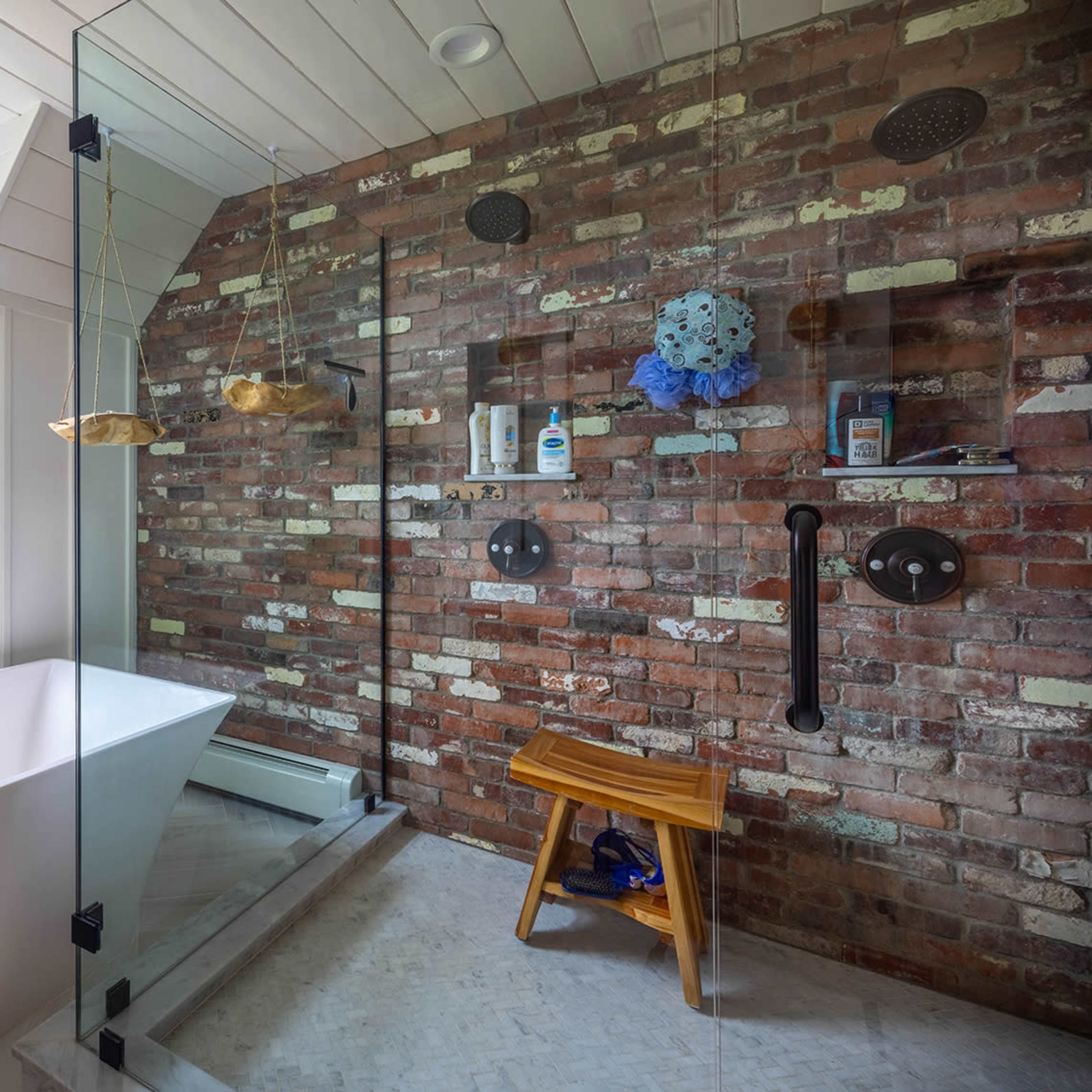 A modern bathroom featuring a walk-in shower with exposed brick walls, a freestanding bathtub, and a large window overlooking a green landscape.