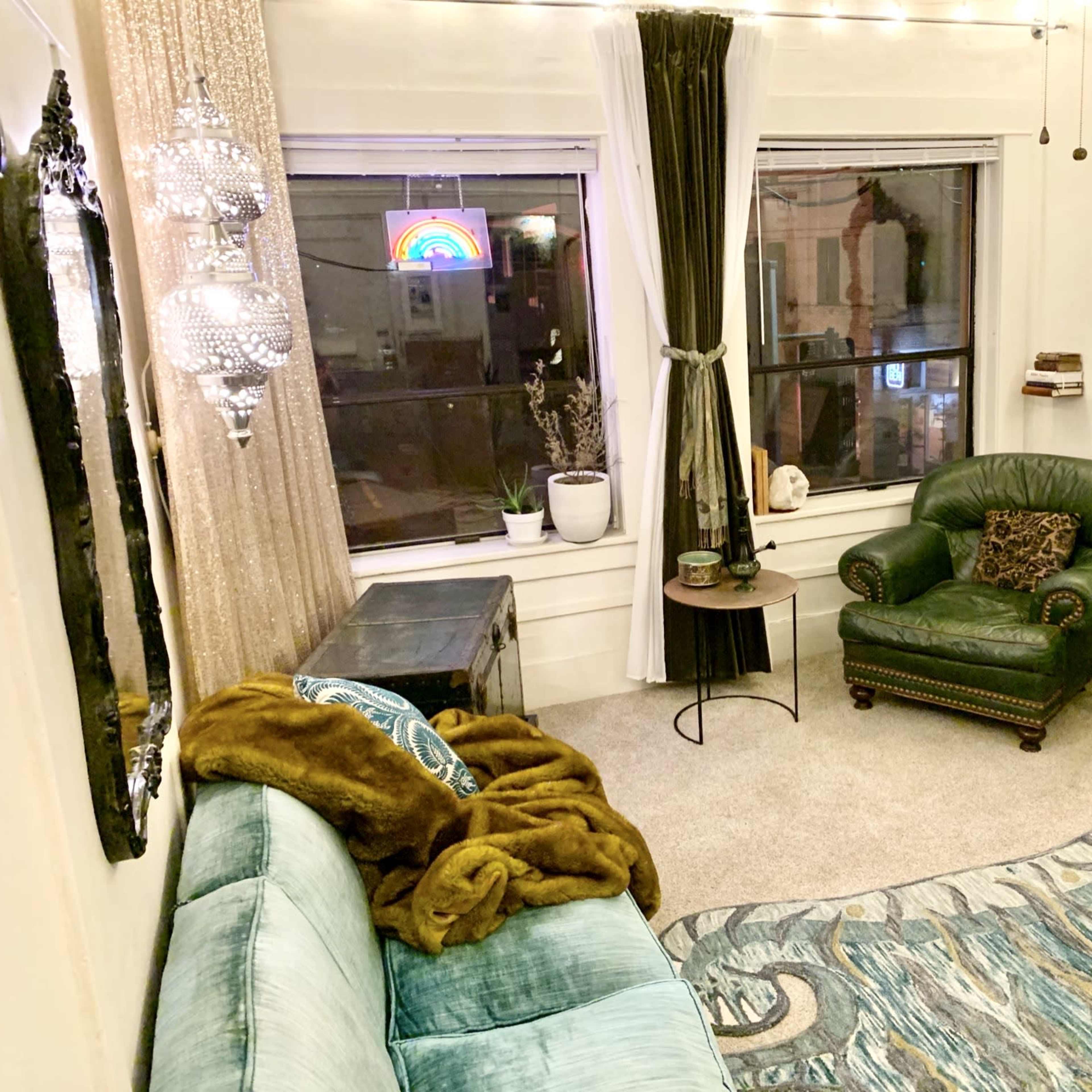 The room features a green armchair, a decorative mirror, a coffee table, a plush rug, and large windows with a colorful neon sign.