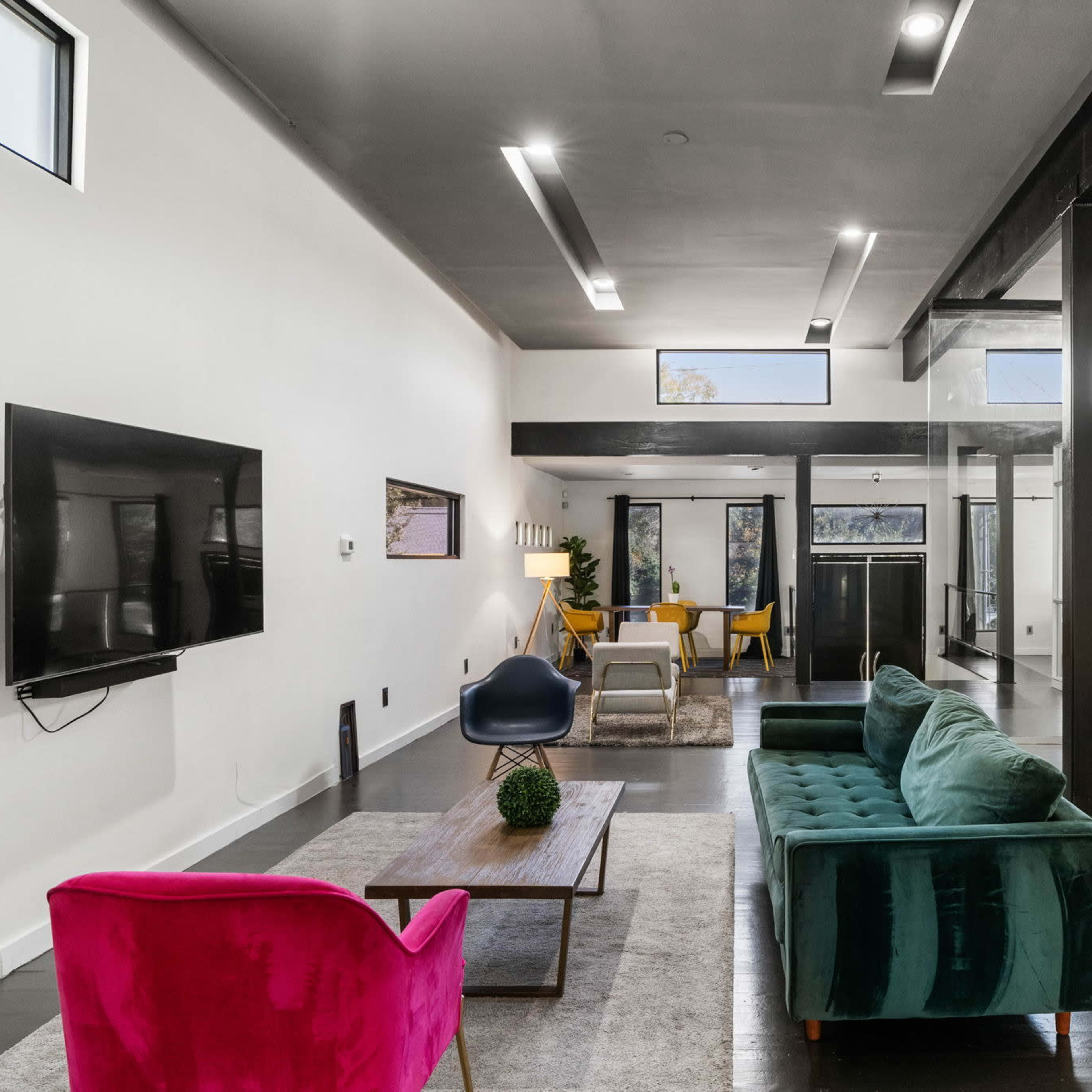 A modern living room with a green couch, a pink chair, a coffee table, a television mounted on the wall, and large windows providing natural light.