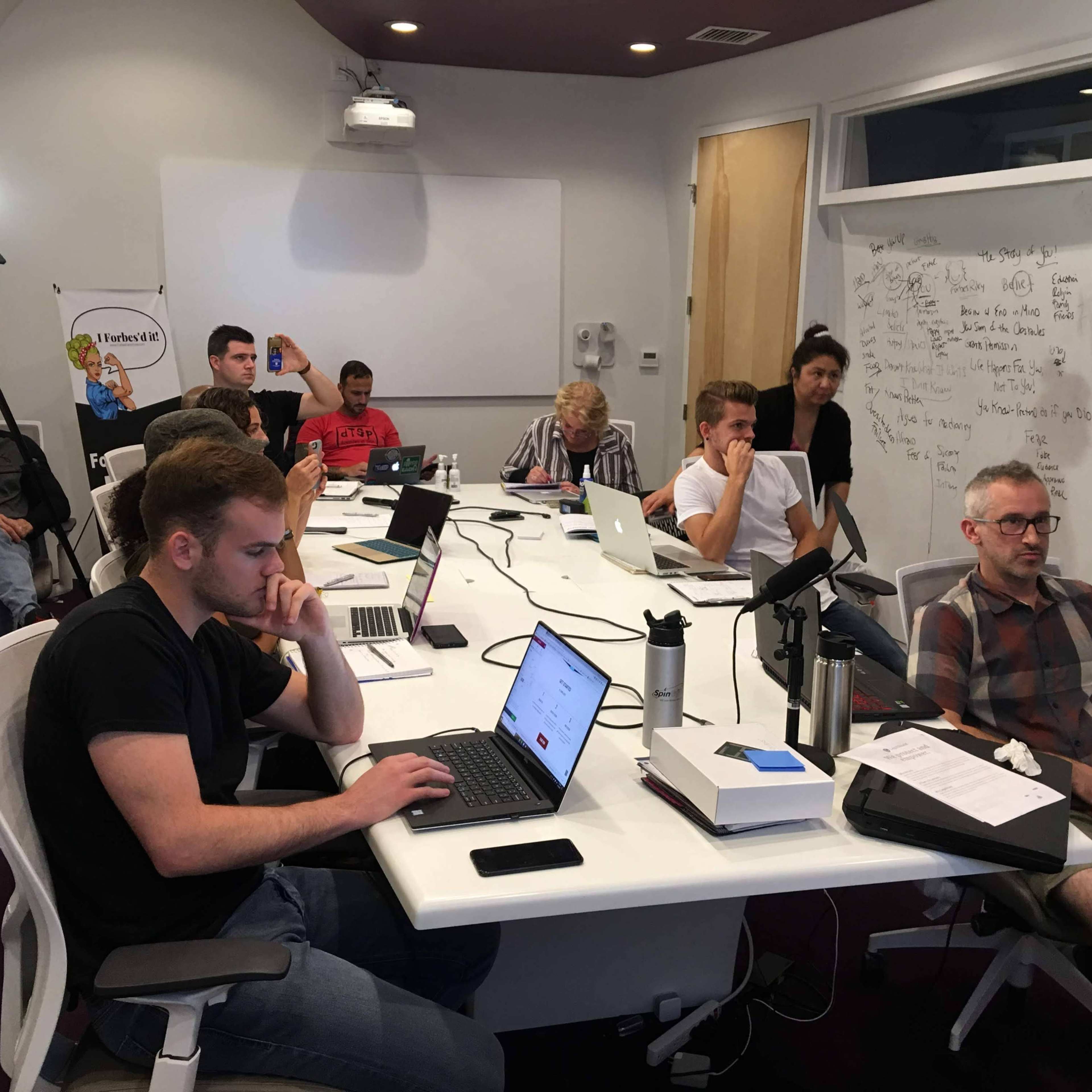 A group of people sits at a long table in a modern conference room, focused on laptops and a whiteboard covered in notes.
