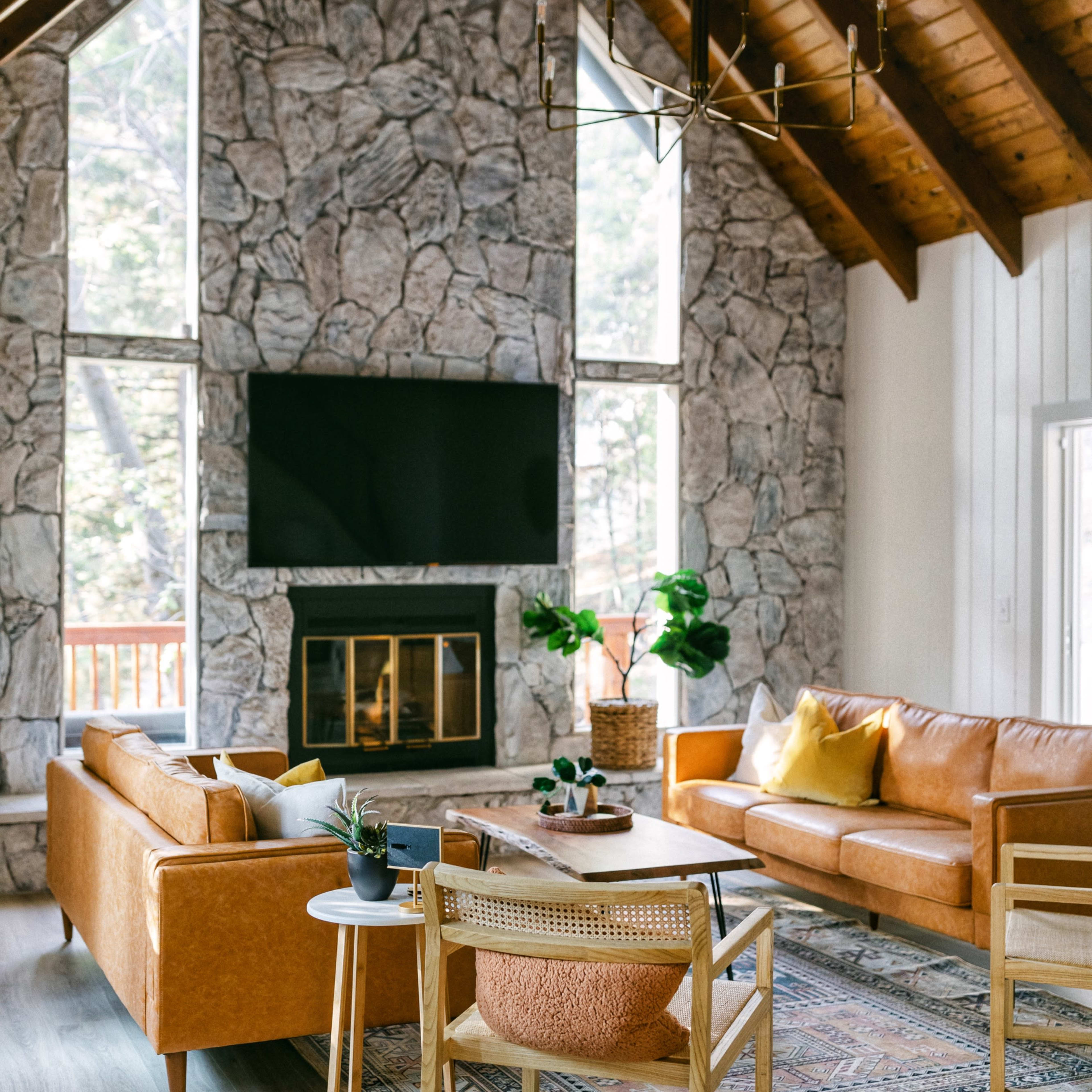 A bright living room with a high stone wall, large windows, a mounted TV, and modern furniture, including leather sofas and a patterned rug.