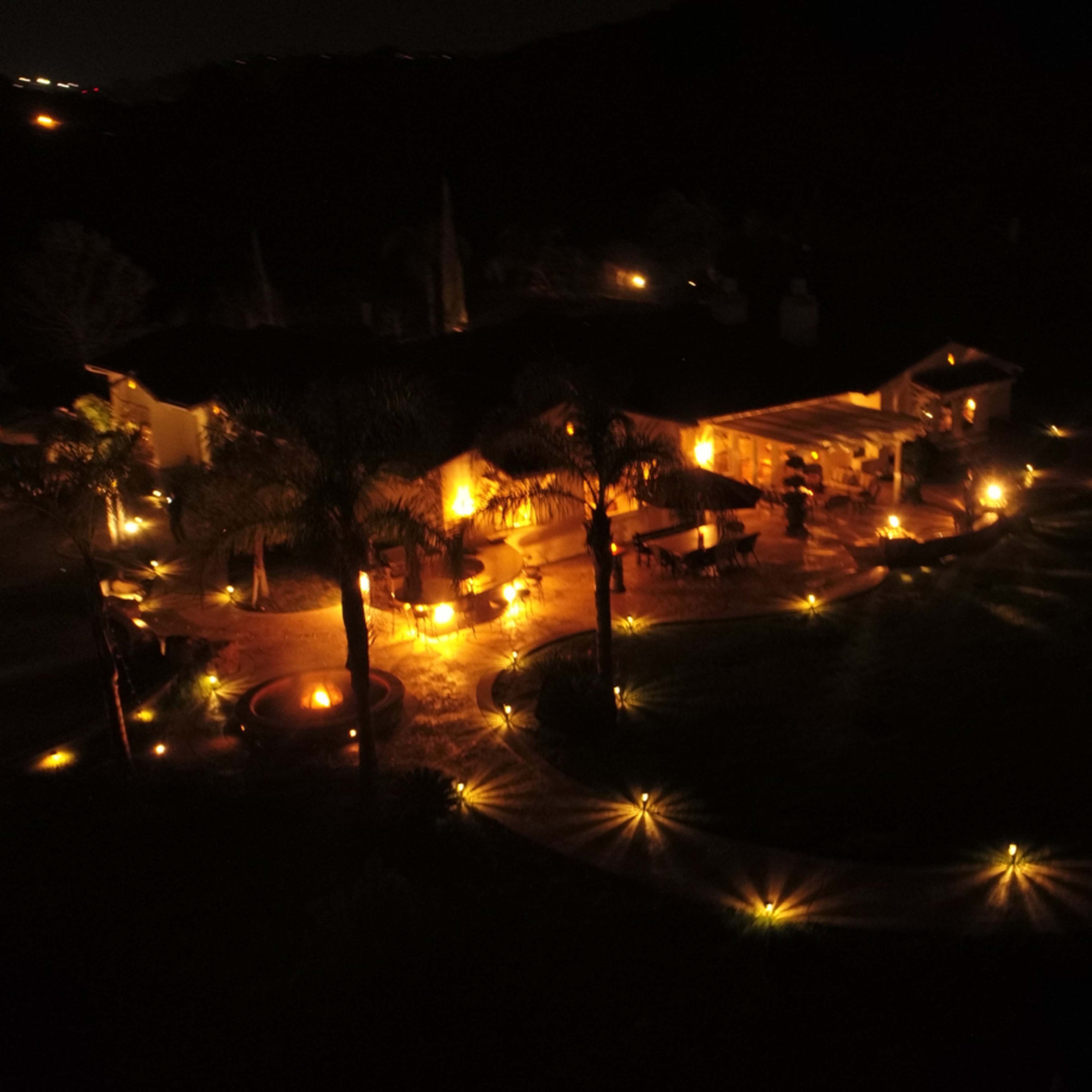 A illuminated outdoor area around a house with palm trees, featuring pathways lined with lights at night.