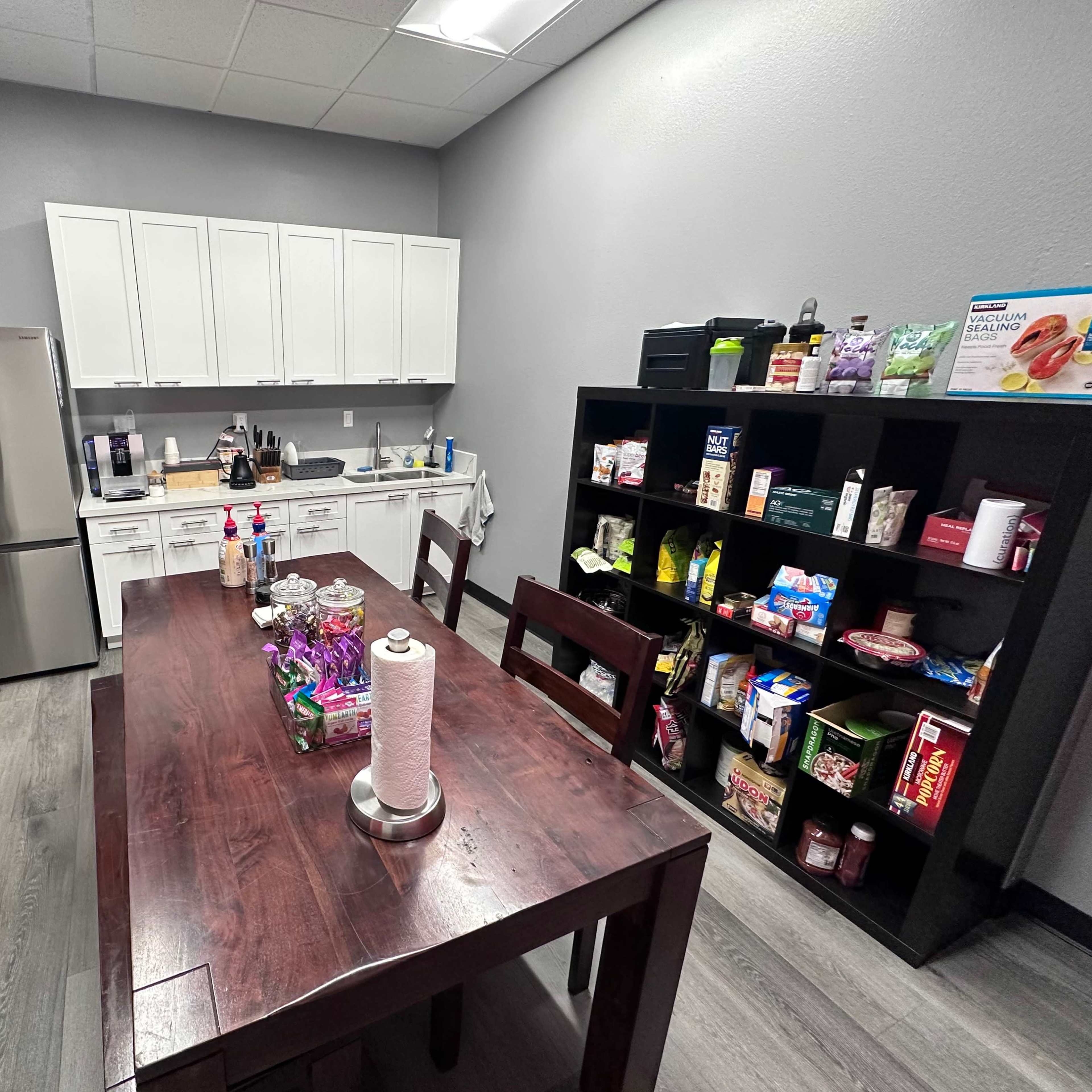 A kitchen area with a dining table, a refrigerator, white cabinets, and a shelf filled with various food items and supplies.