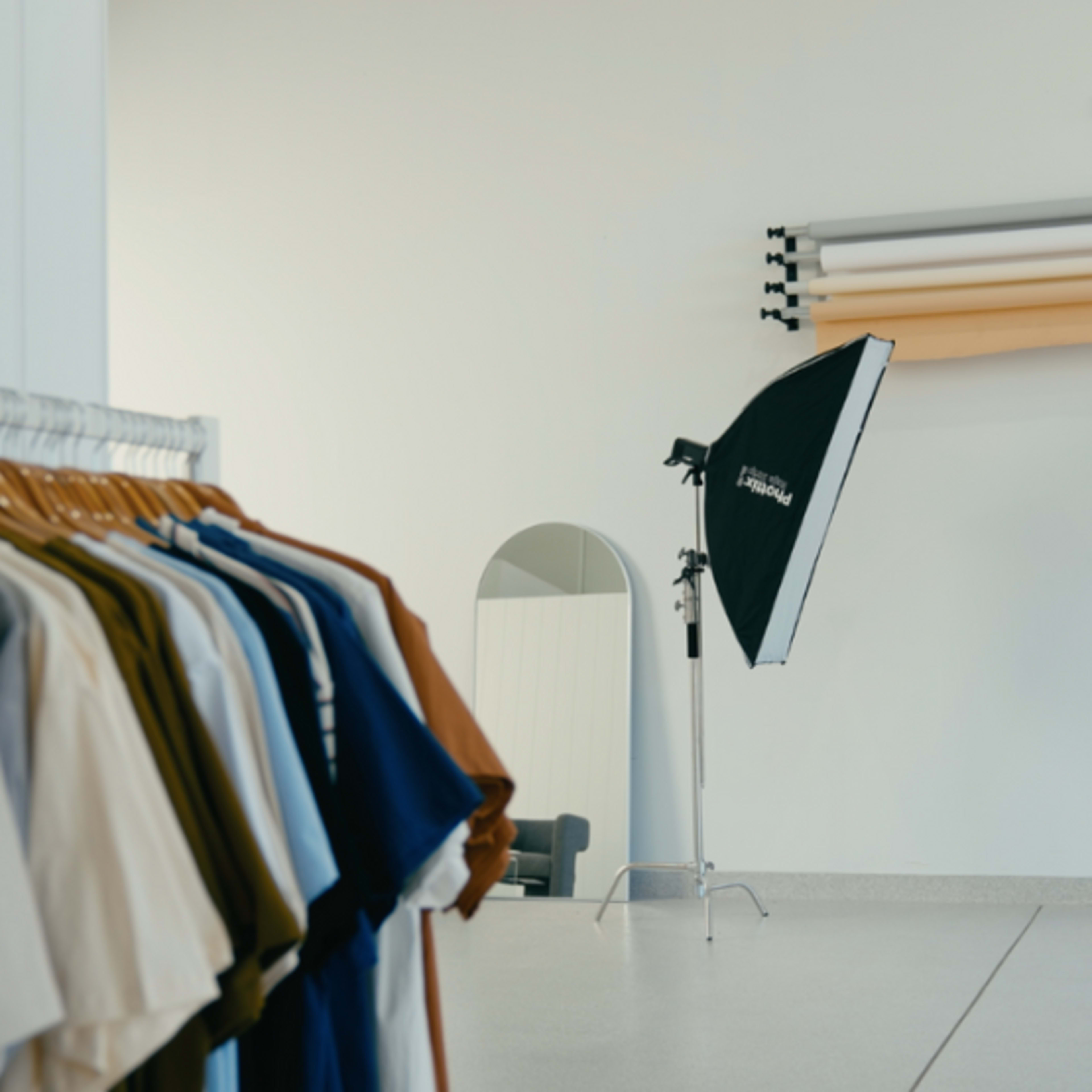 A clothing rack displays various colored shirts next to a photography setup featuring a backdrop and studio lights in a minimalist room.