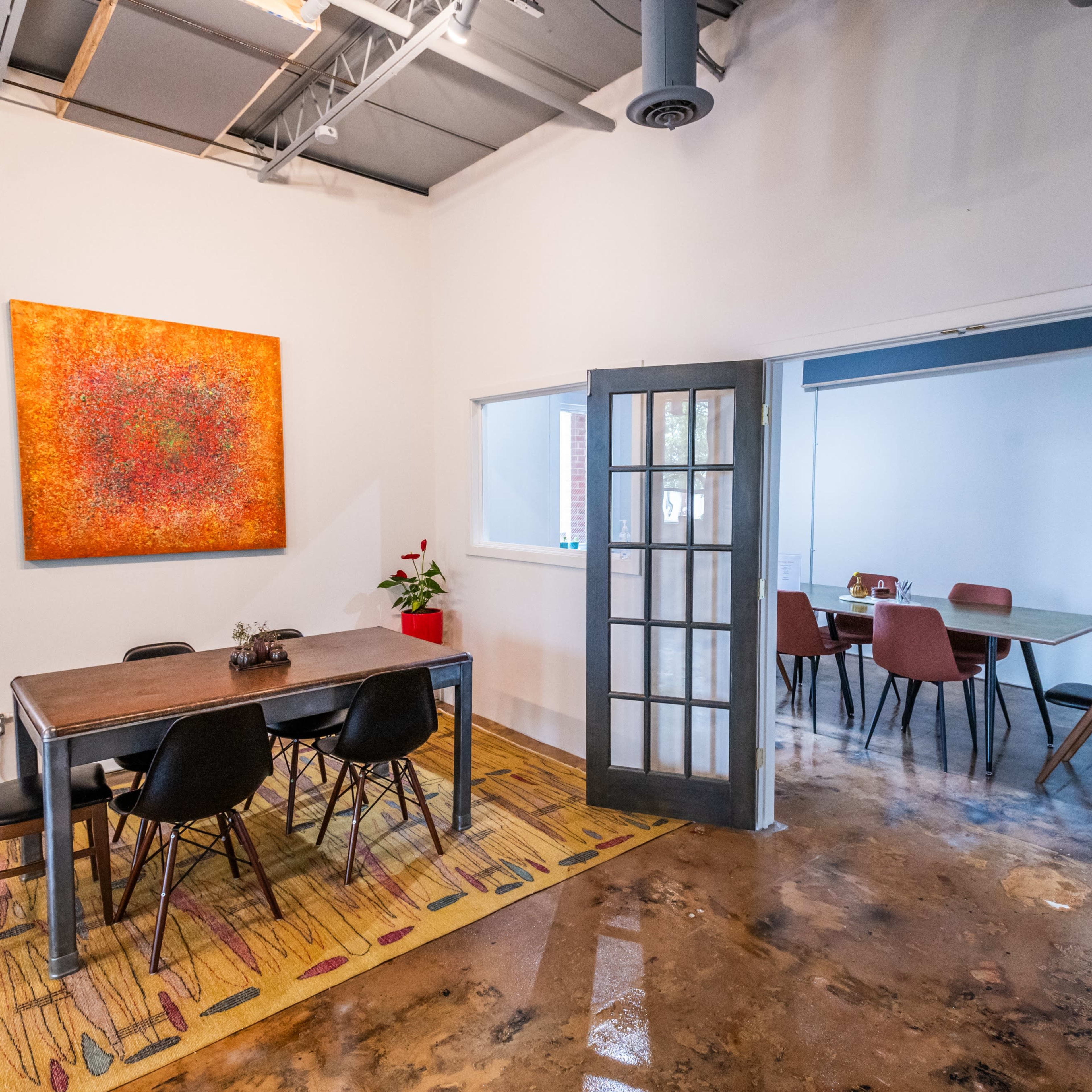 A modern meeting space with a large wooden table surrounded by black chairs, an orange artwork on the wall, and a second room visible through glass doors featuring additional seating.