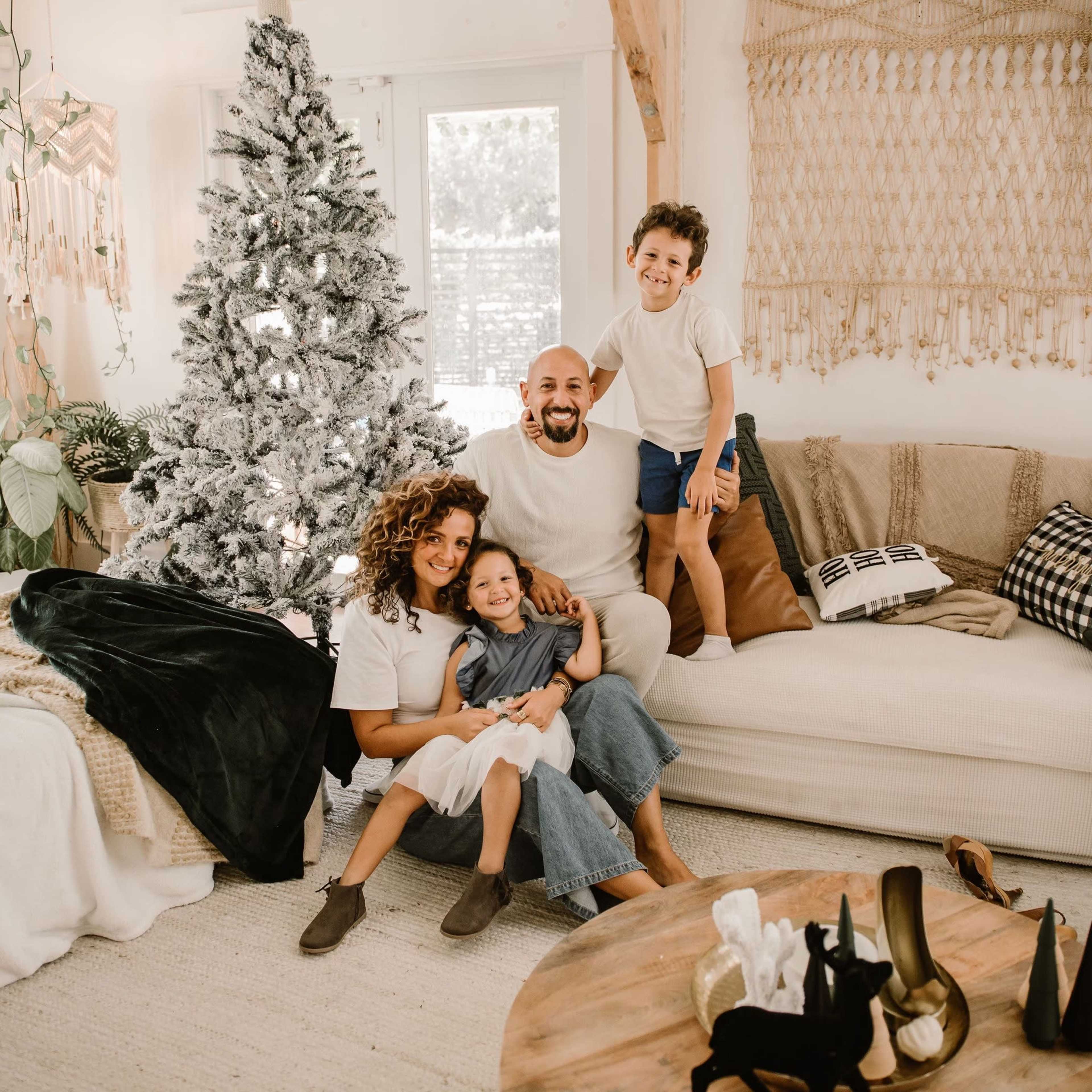 A family of four, including two children, poses together in a cozy living room decorated with a Christmas tree and warm textiles.