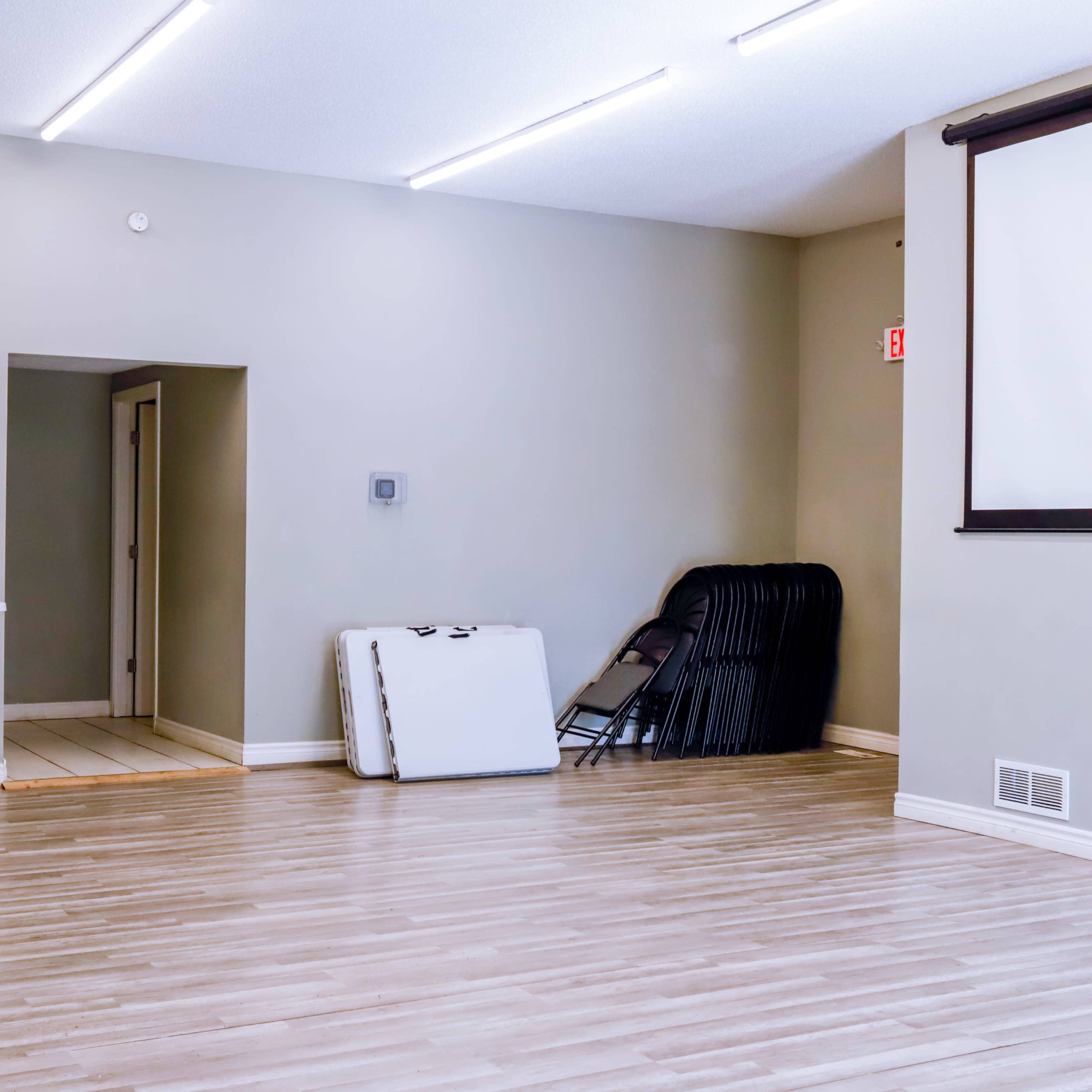 A spacious room with hardwood flooring, featuring a projector screen on one wall, a table with a laptop, and stacked folding chairs in the corner.