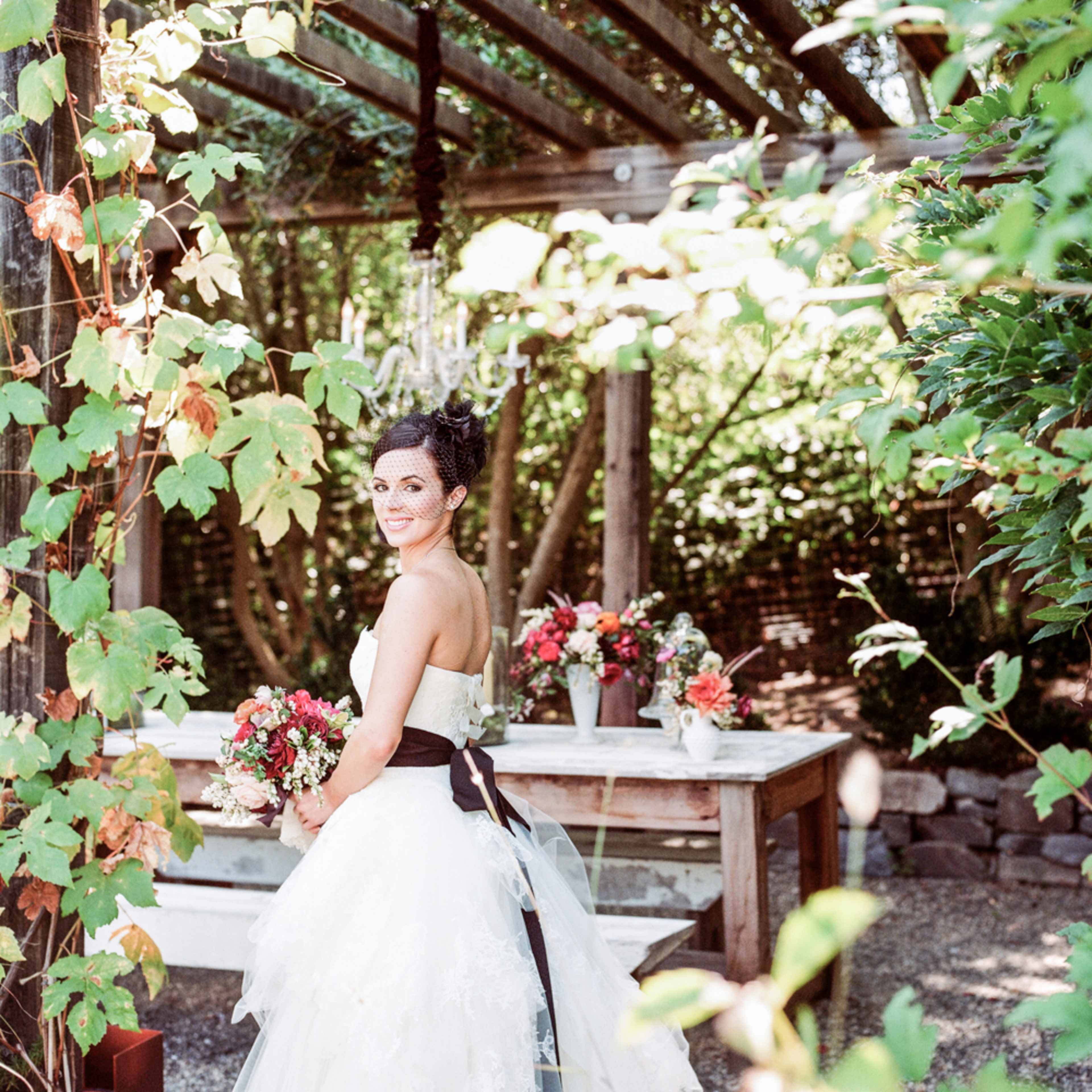 A bride in a strapless wedding gown holds a bouquet and stands near a decorated table under a trellis adorned with greenery.