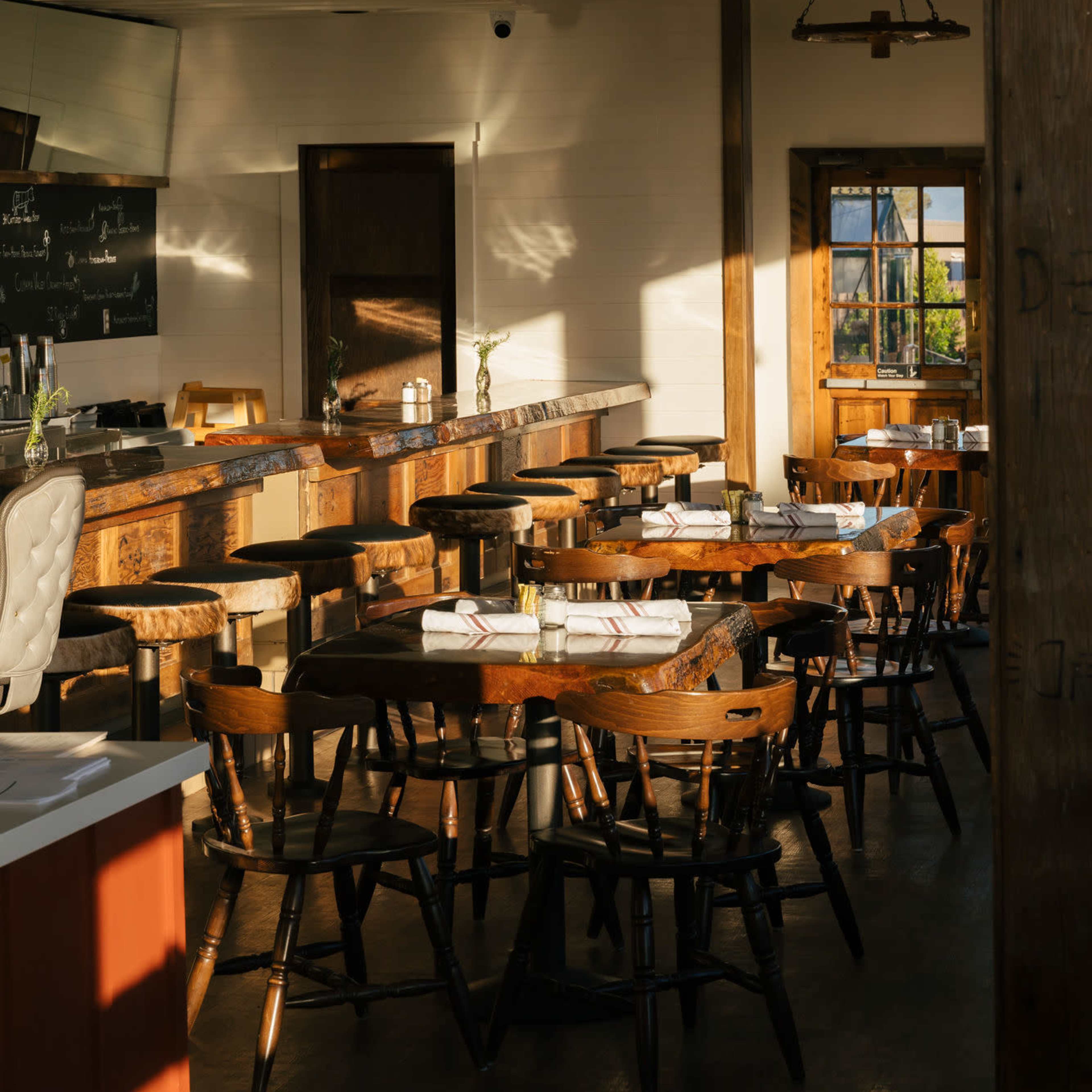 A rustic restaurant interior with wooden tables and chairs, a long bar, and large windows allowing natural light to enter.