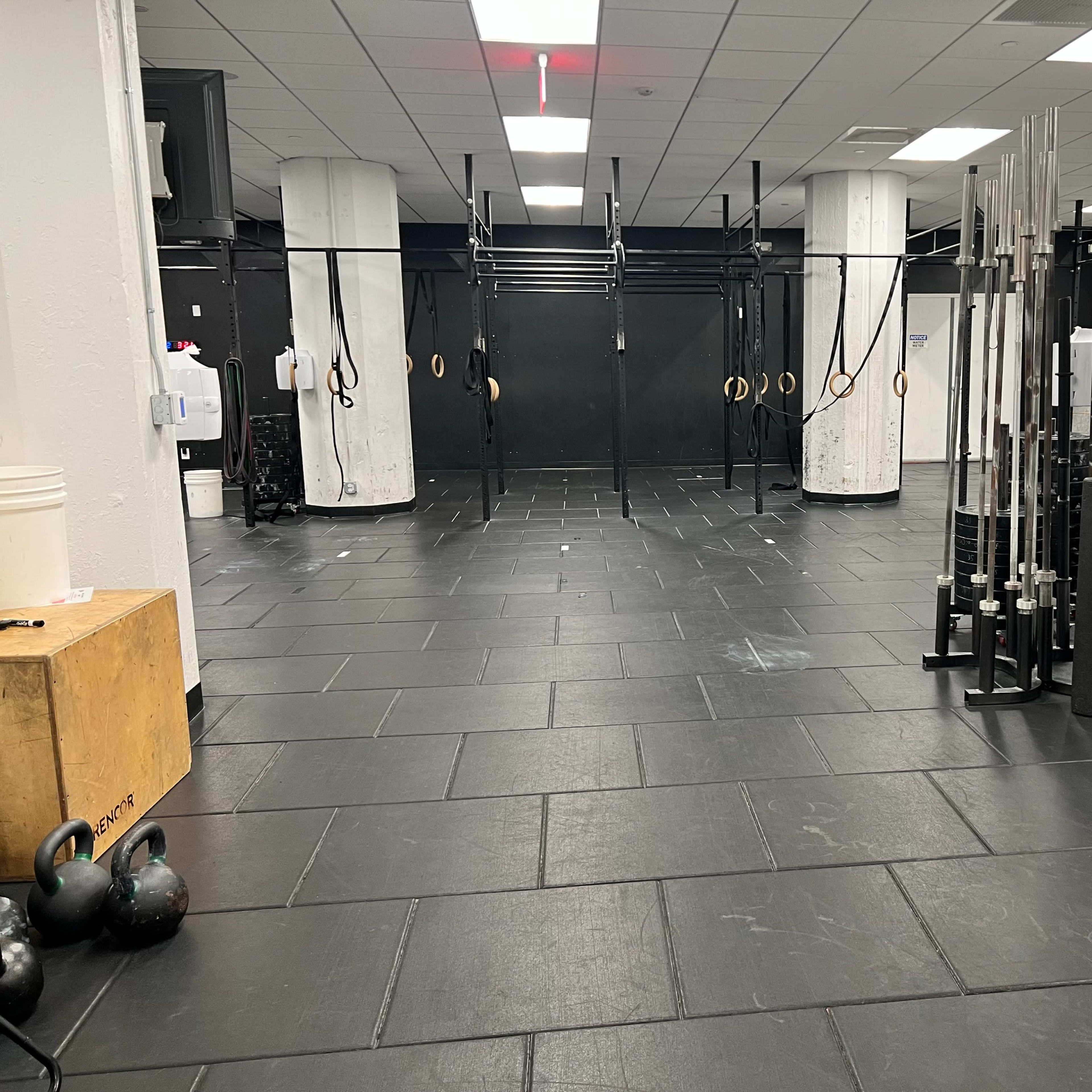 A spacious gym area with various exercise equipment, including kettlebells, a wooden box, and weightlifting bars, arranged on black rubber flooring.