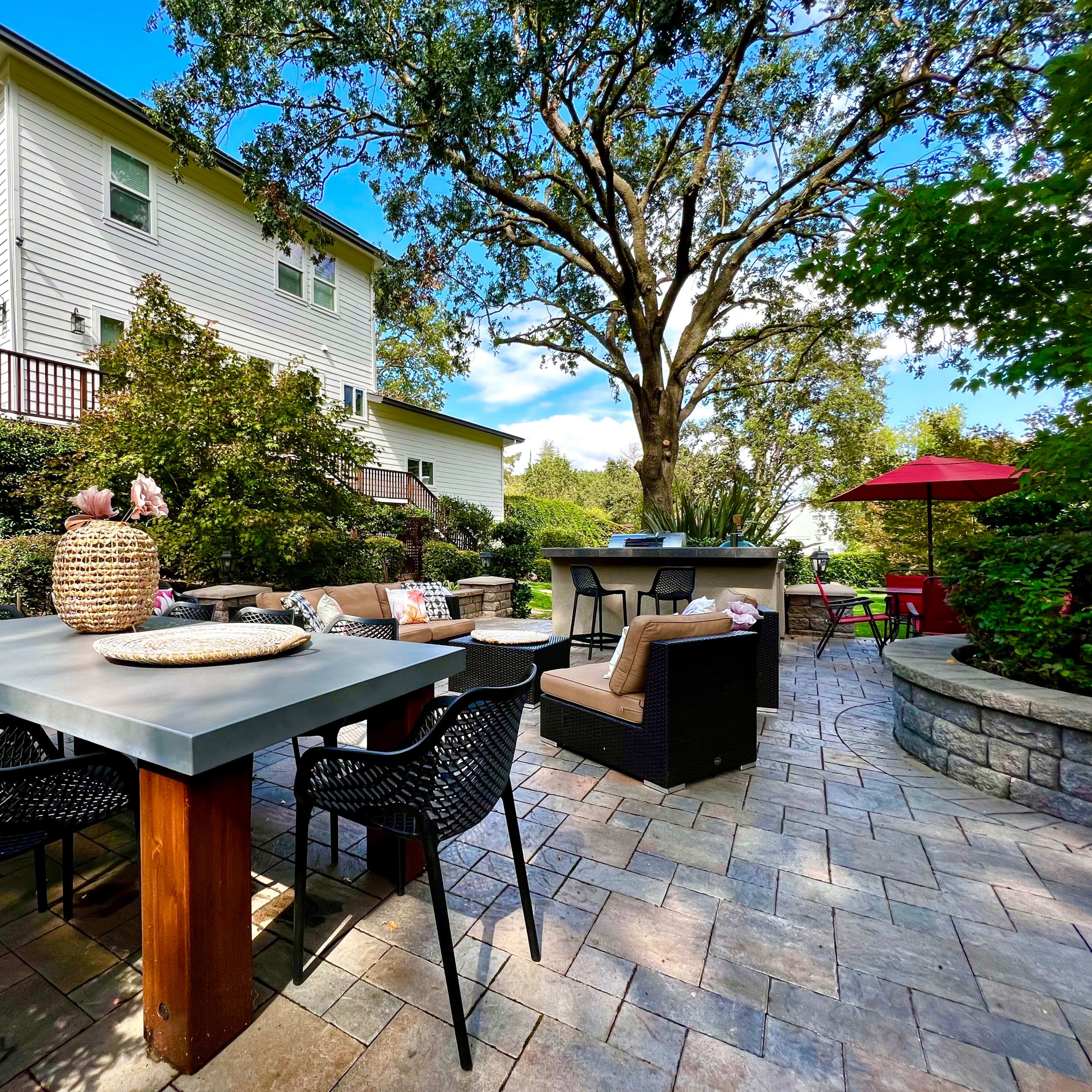The scene shows a patio area with a large table, chairs, and a barbecue station, surrounded by greenery and a white house in the background.