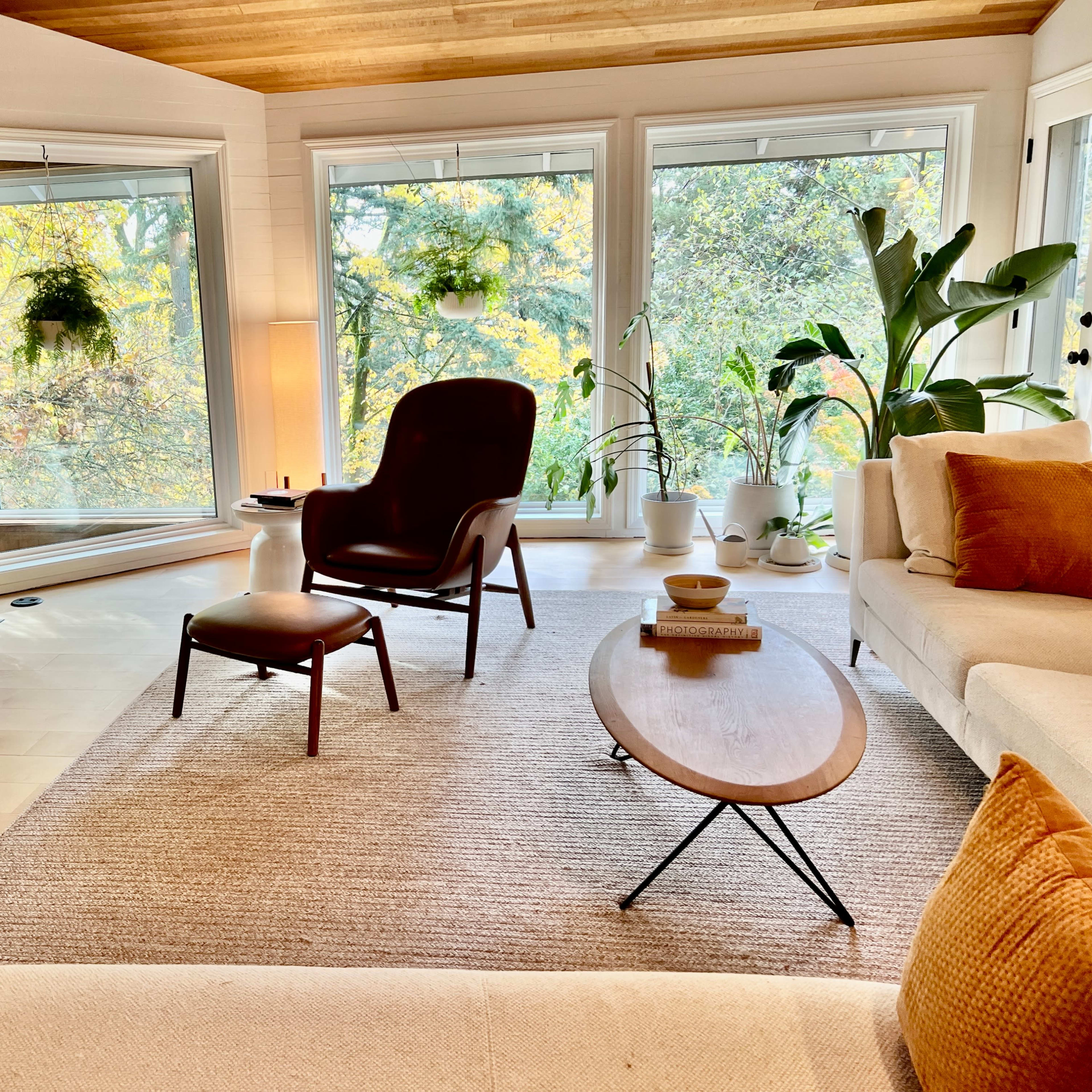 A modern living room features a comfortable armchair and ottoman, a coffee table, and large windows showcasing greenery outside.
