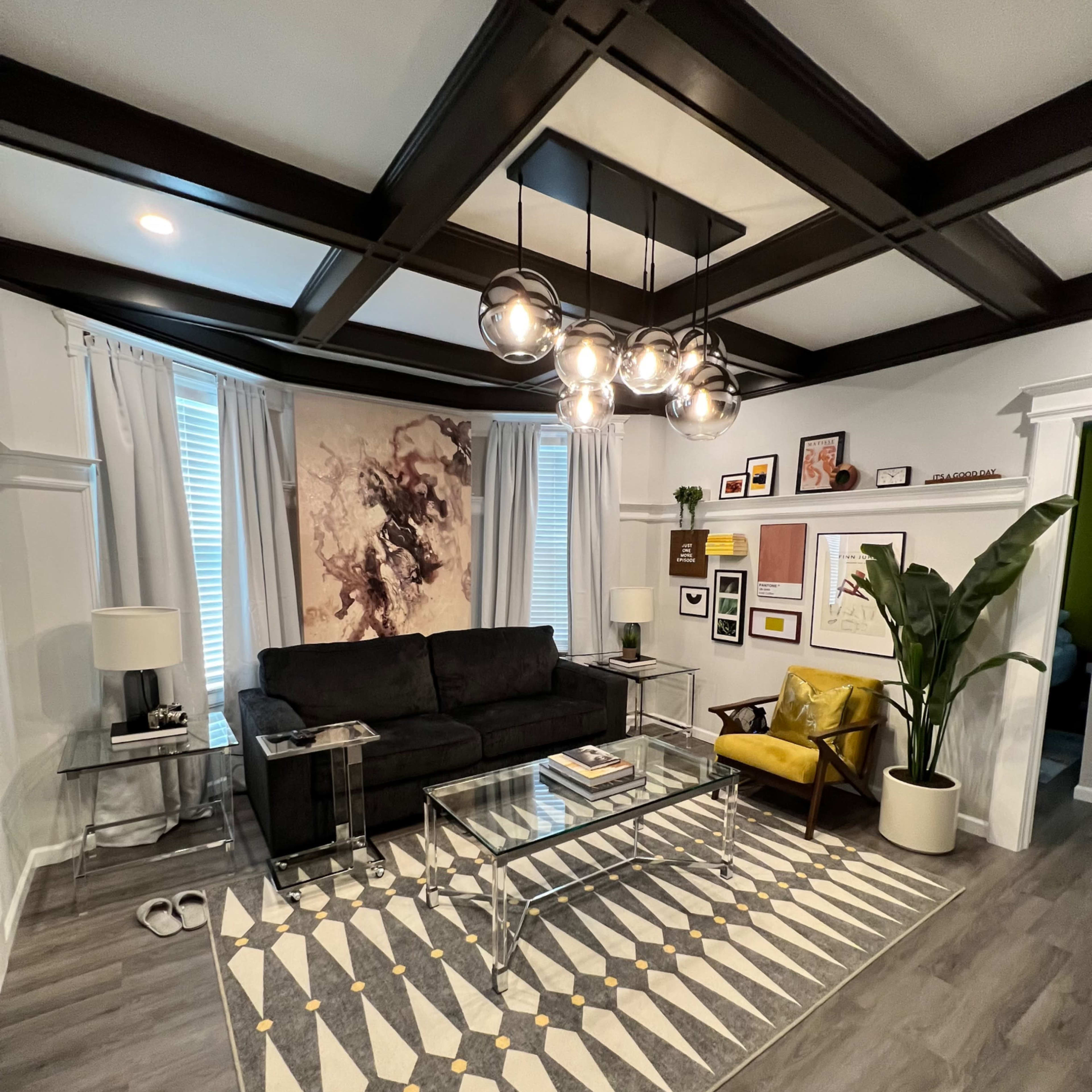 A modern living room featuring a black couch, a yellow armchair, a large wall art piece, and a coffee table on a patterned rug, with decorative plants and framed photographs on the walls.