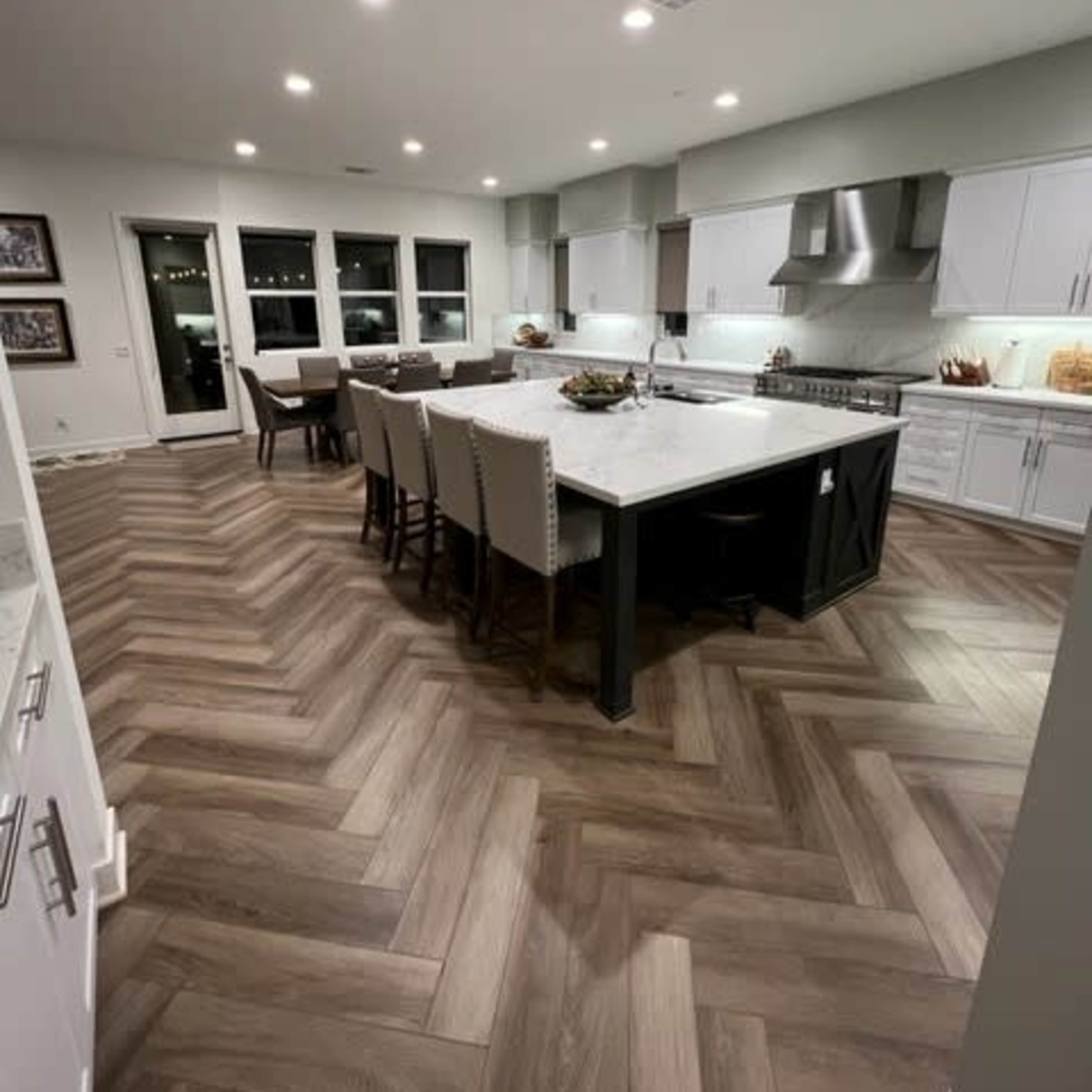 A modern kitchen with a large island, seating for six around it, white cabinetry, and herringbone-patterned wooden flooring.