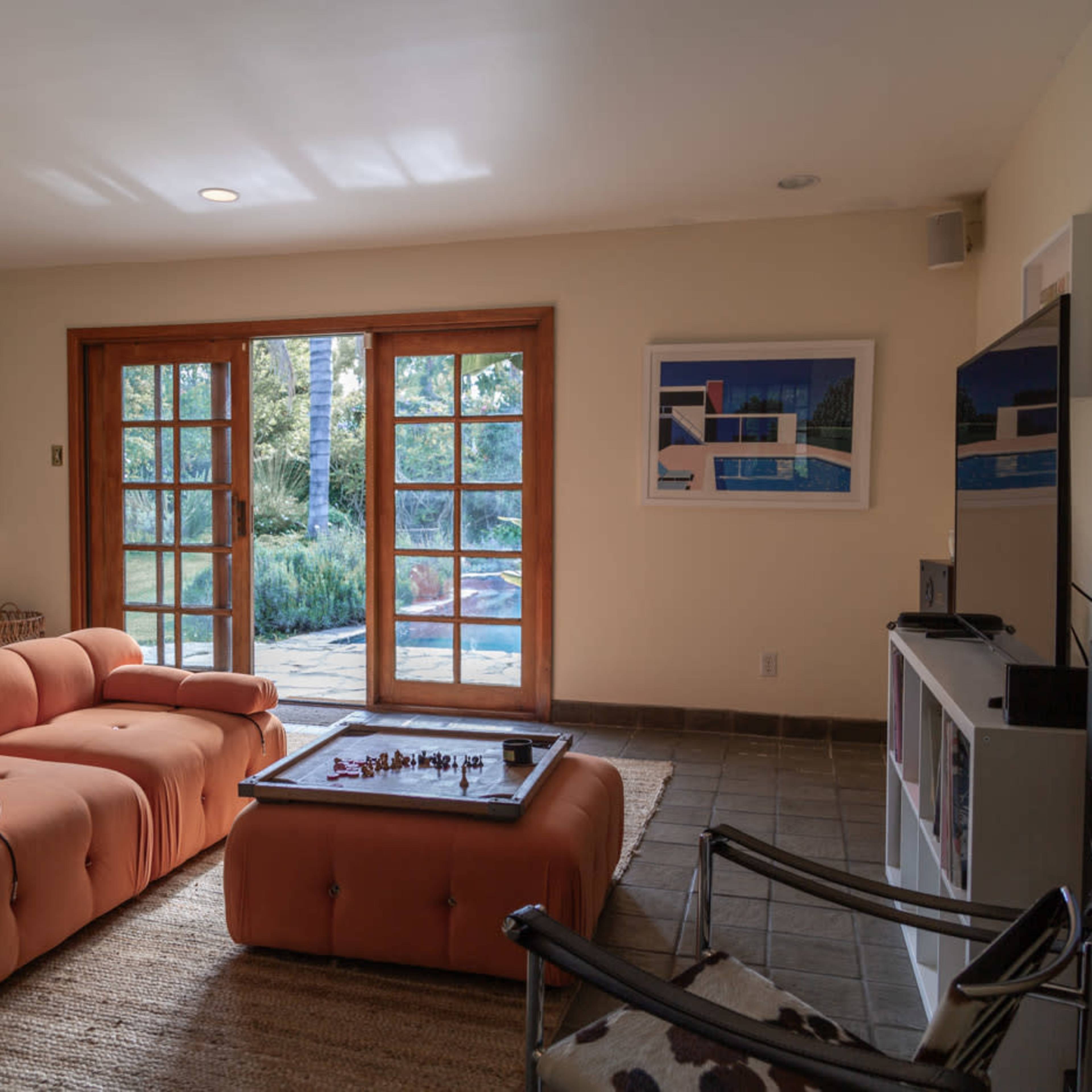 A bright living room features a peach-colored sofa and a glass-top coffee table with a chess set, set against a backdrop of large double doors leading to a patio.